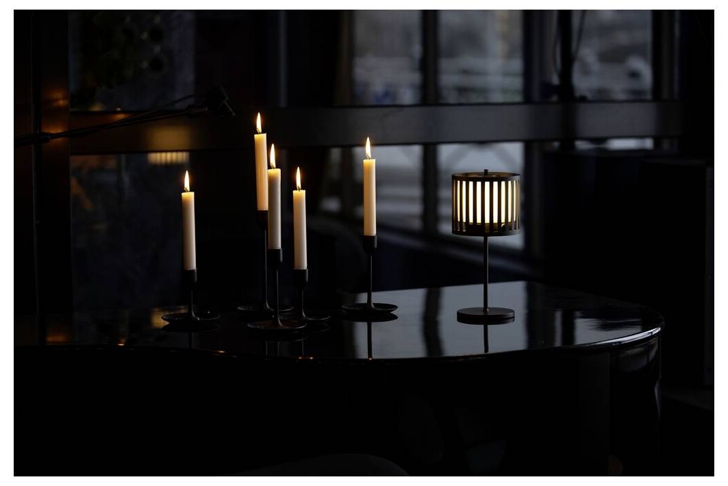 Candele accese su un pianoforte scuro, che riflettono una luce soffusa; una lampada moderna aggiunge un bagliore ambientale, in una stanza scarsamente illuminata con grandi finestre.