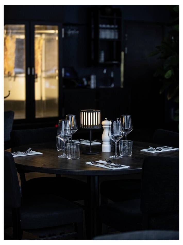 Un tavolo di un ristorante scarsamente illuminato, apparecchiato per quattro persone, con bicchieri da vino, bicchieri d'acqua, posate e una lampada decorativa accesa al centro.