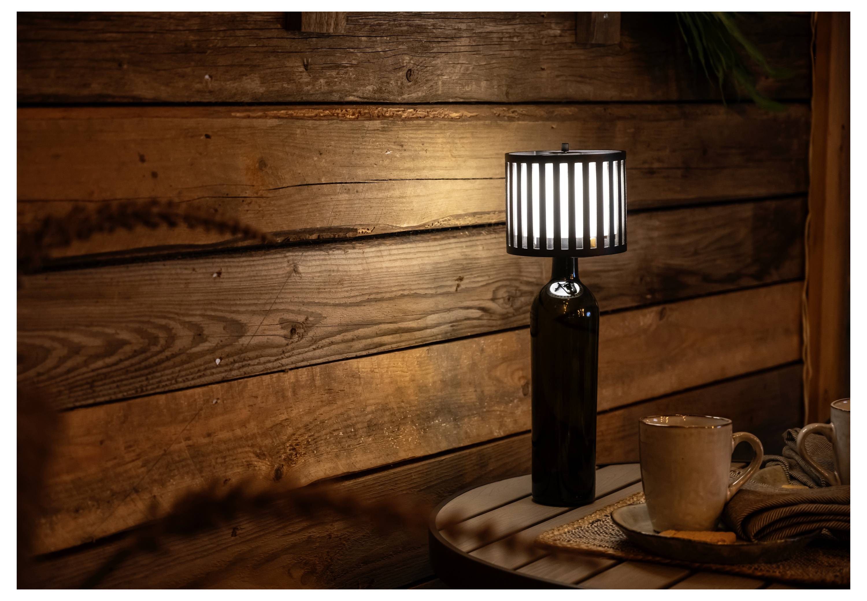 Una stanza accogliente in legno presenta una lampada unica realizzata con una bottiglia di vetro con un paralume a righe su un tavolo rotondo, accanto a tazze di caffè e grano.