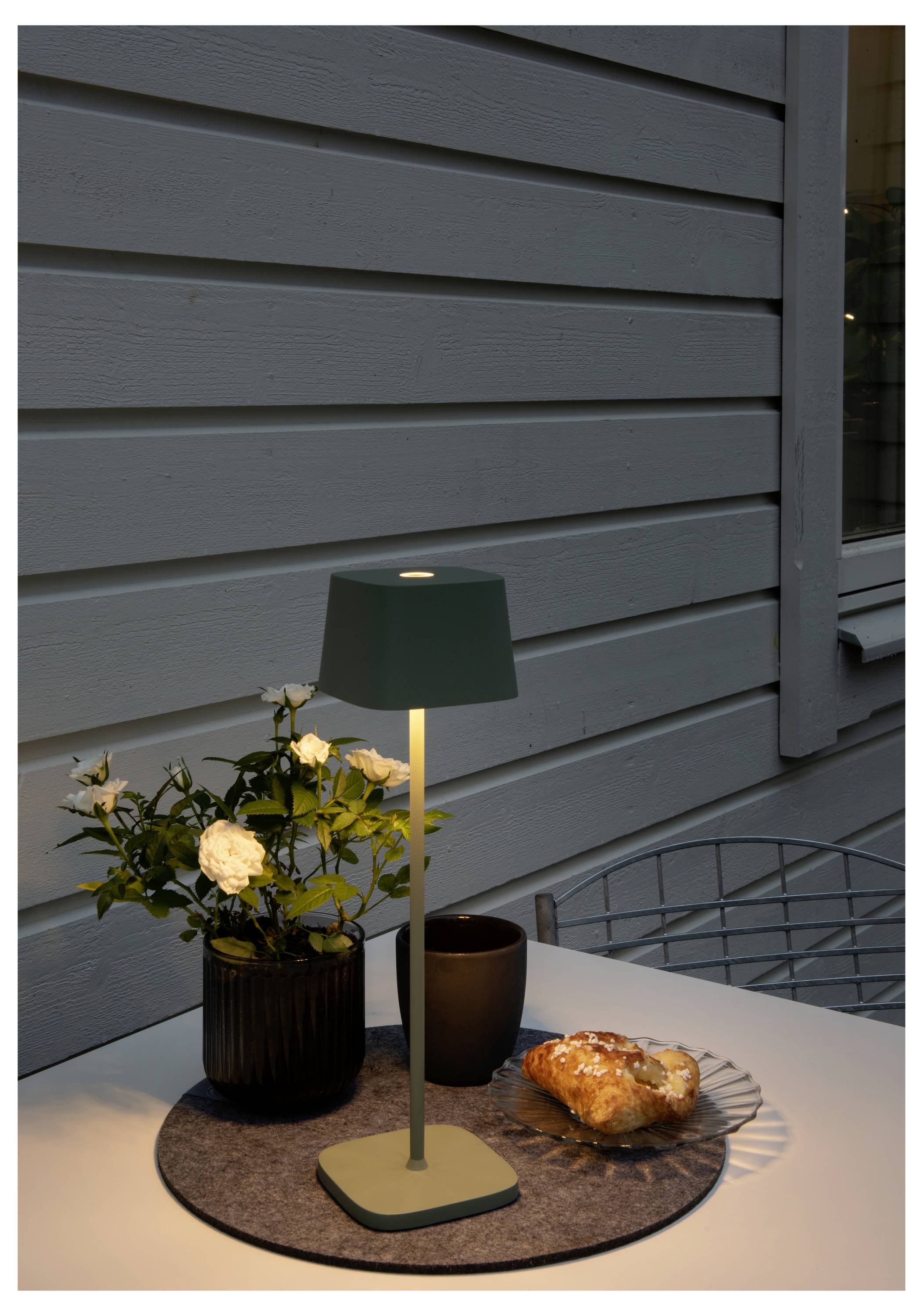 Un accogliente allestimento di un tavolo all'aperto che presenta una lampada moderna, un vaso con fiori bianchi, un croissant su un piatto e una tazza scura.