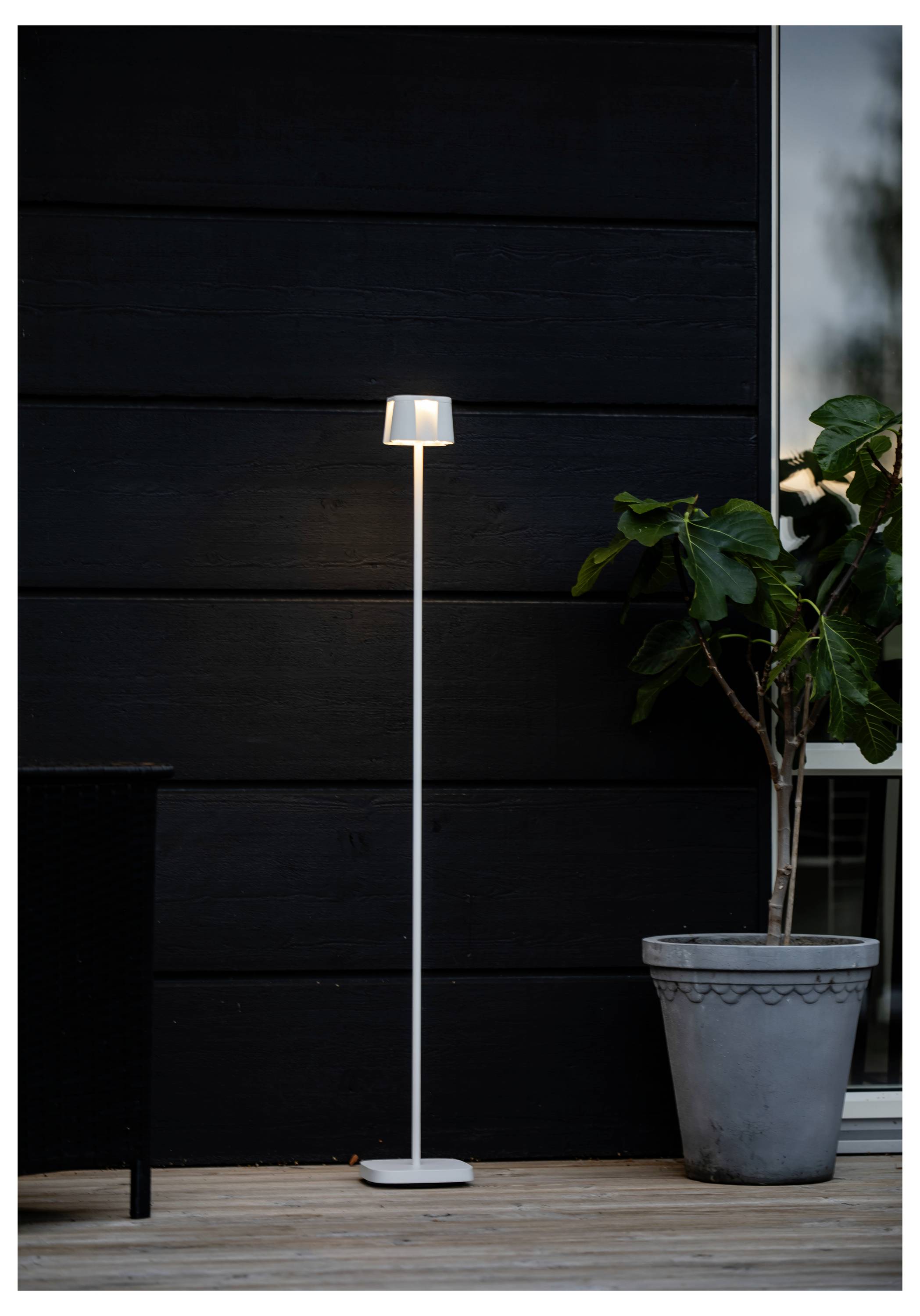 Una lampada da terra alta e sottile con un paralume geometrico è accesa contro un muro esterno scuro, accanto a una grande pianta in vaso su una terrazza in legno.