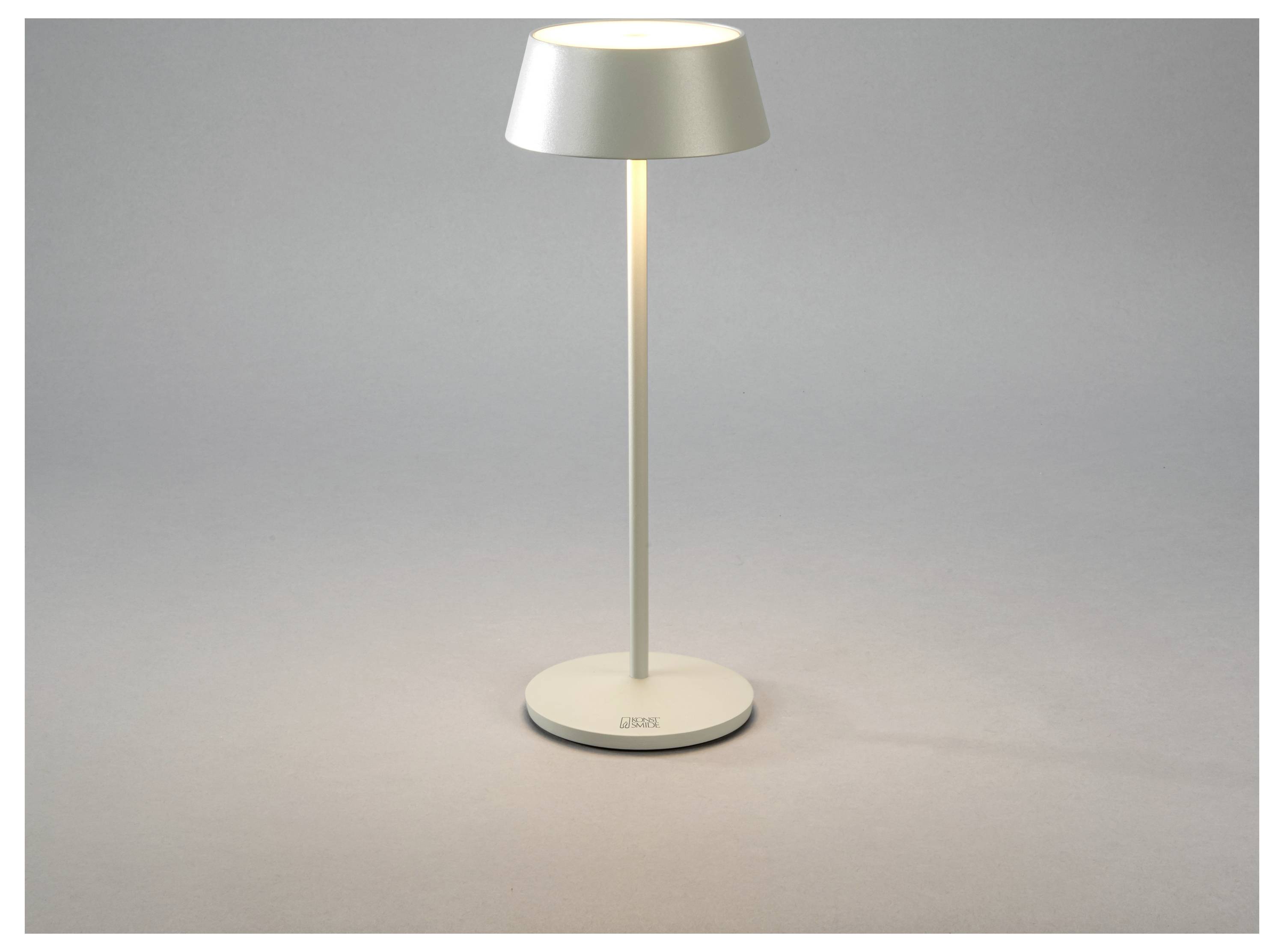 Una lampada da tavolo moderna, dal tono argentato, con paralume conico e base sottile, poggia su una superficie grigia, emettendo una luce calda.