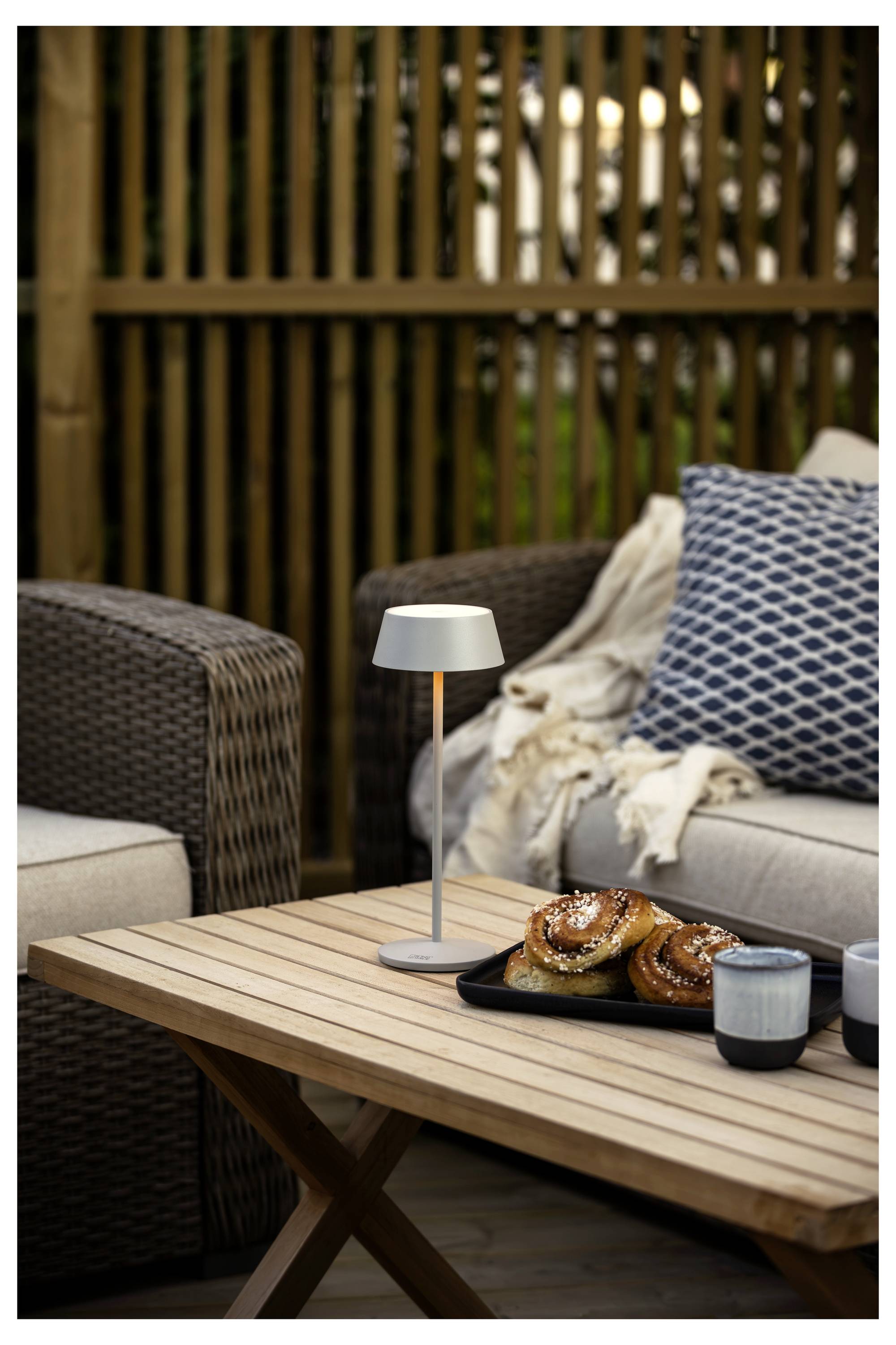 Un'area per sedere all'aperto con mobili in vimini presenta un tavolo di legno con un piatto di pasticcini, tazze e una piccola lampada. Una coperta accogliente e un cuscino sono visibili.