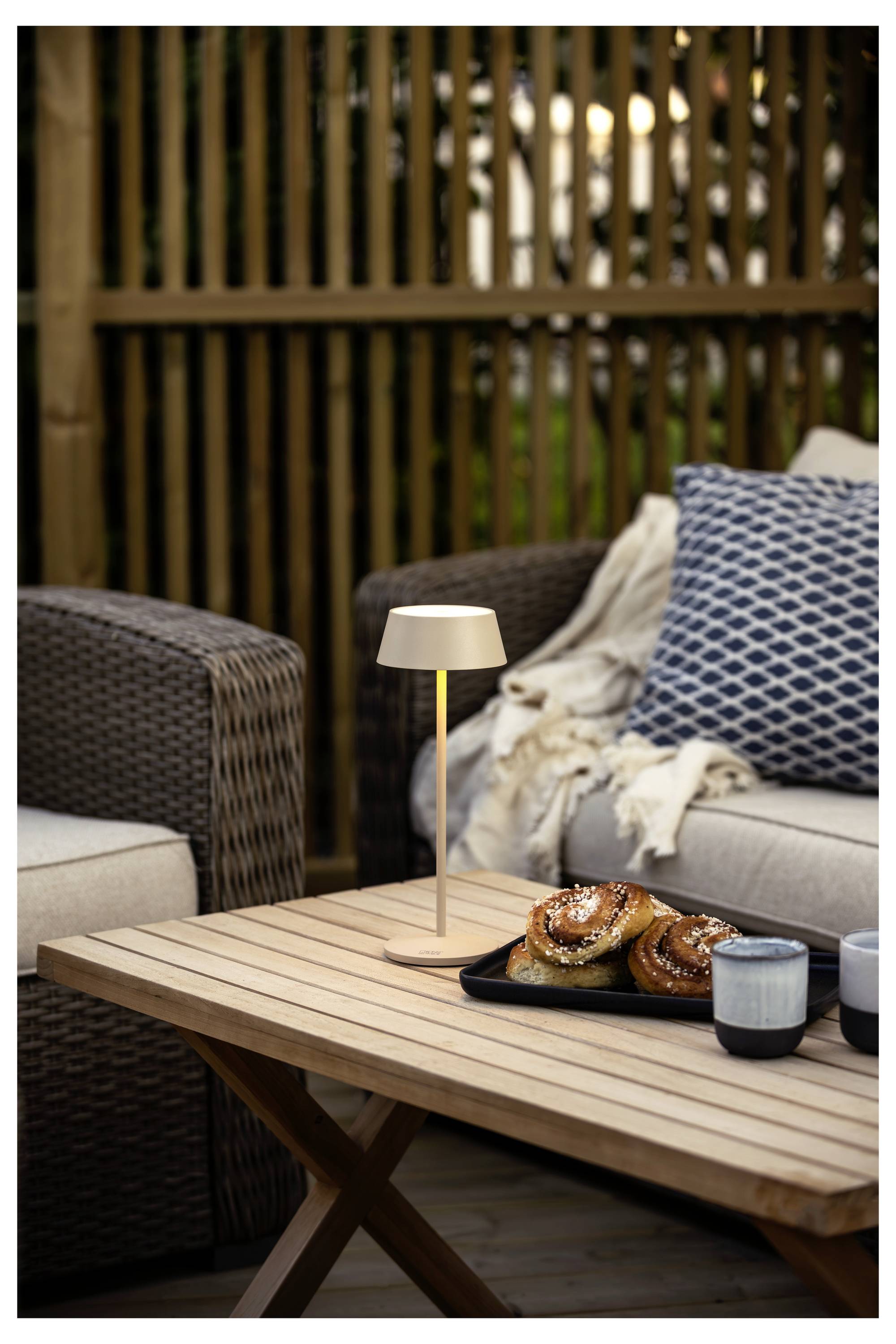 Un accogliente patio esterno con un tavolo di legno che regge un vassoio di pasticcini e due tazze. Un divano con cuscini e una coperta sullo sfondo.