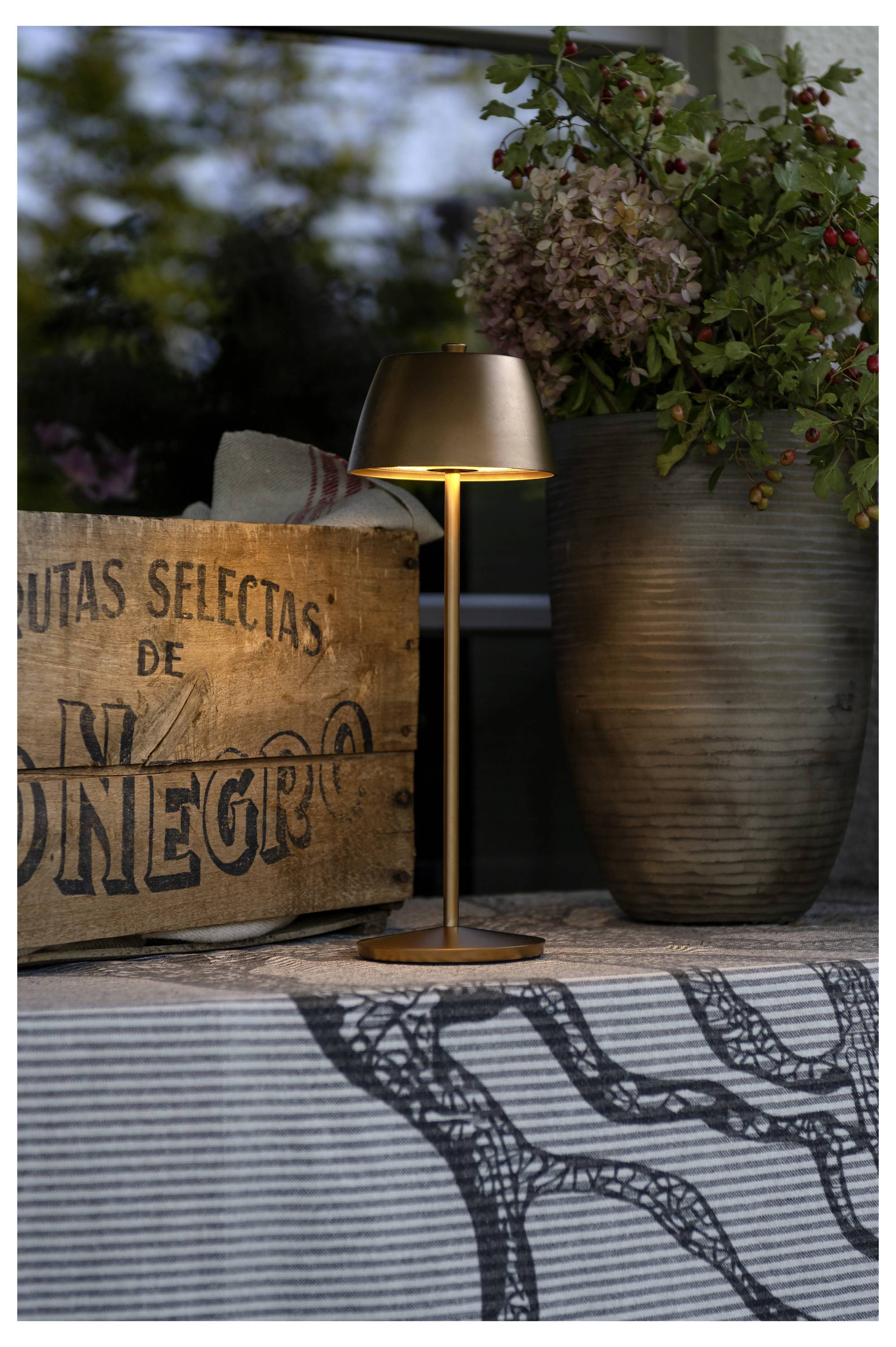 Una lampada da tavolo accesa con finitura metallica è posizionata su un tavolo coperto da un panno, accanto a una scatola di legno rustica e un vaso con fiori.