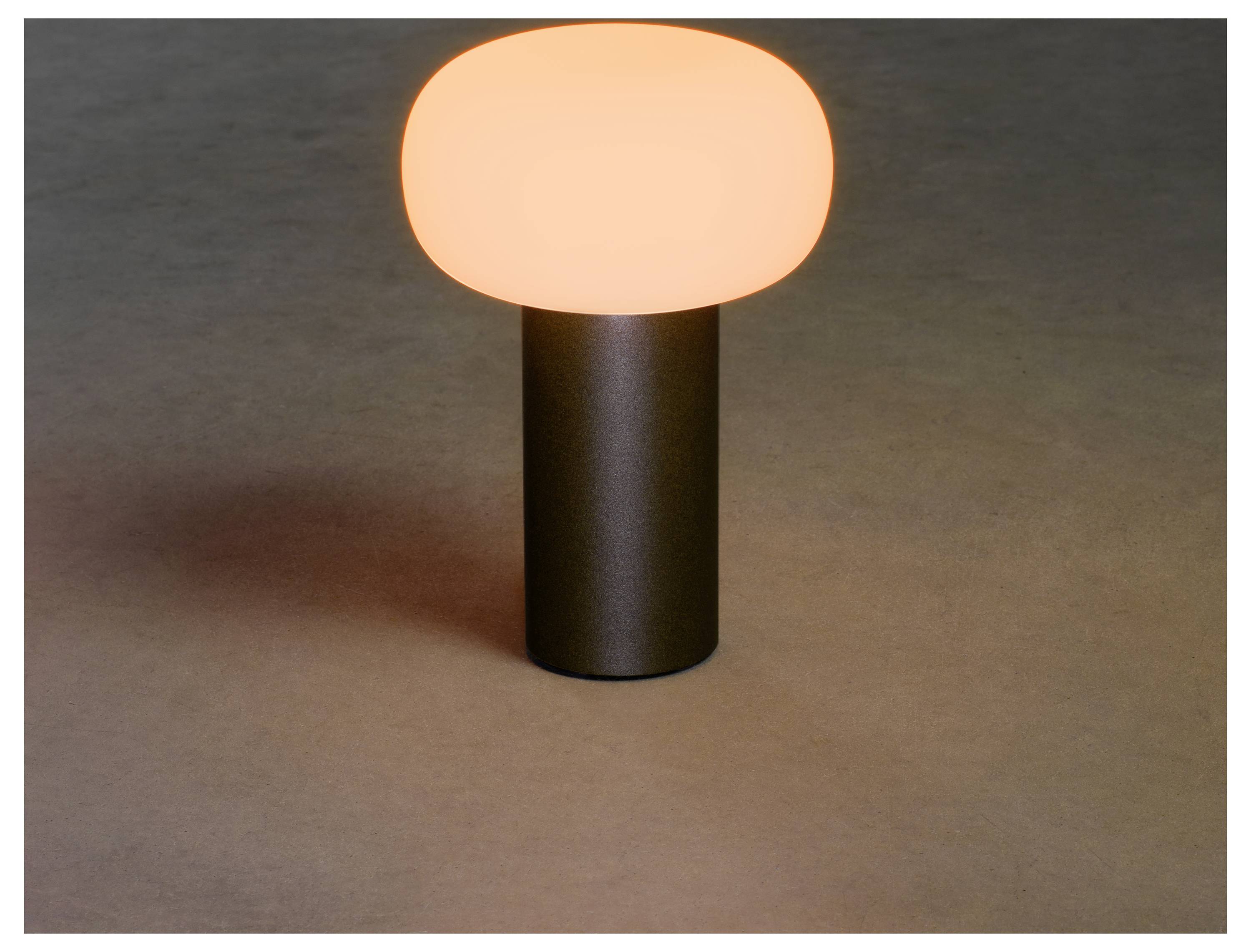 Una piccola lampada da tavolo moderna con un paralume ovale luminoso è posizionata su una superficie piana, diffondendo una luce calda.