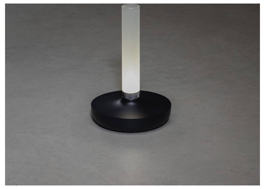 Una lampada moderna, cilindrica, bianca con una luce che emana un bagliore, poggiata su una base rotonda scura, posizionata su una superficie grigia liscia.