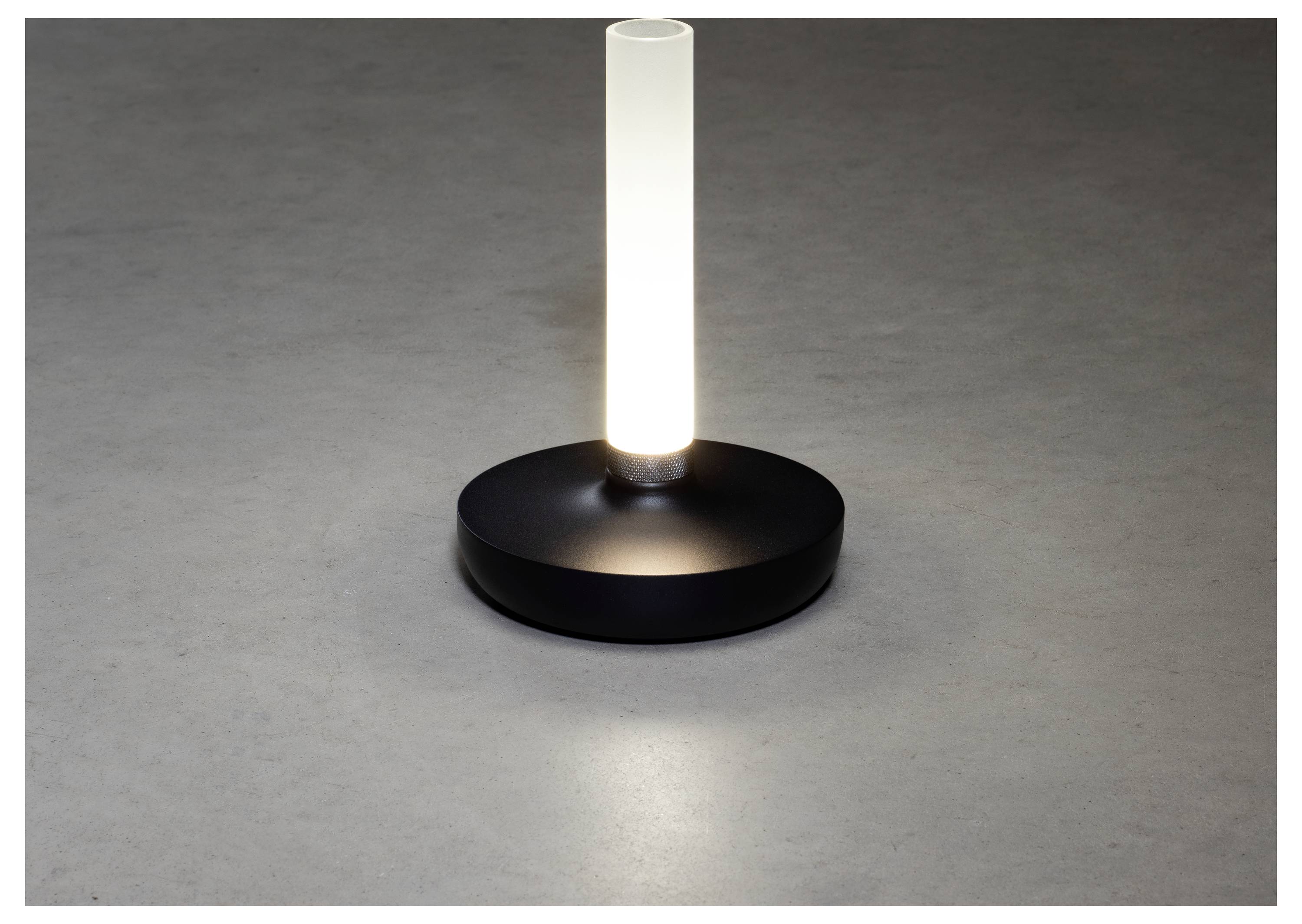 Una lampada da tavolo moderna, cilindrica, emette una luce bianca soffusa da una colonna di vetro smerigliato, poggiata su una base circolare semplice e nera, contro una superficie grigia.