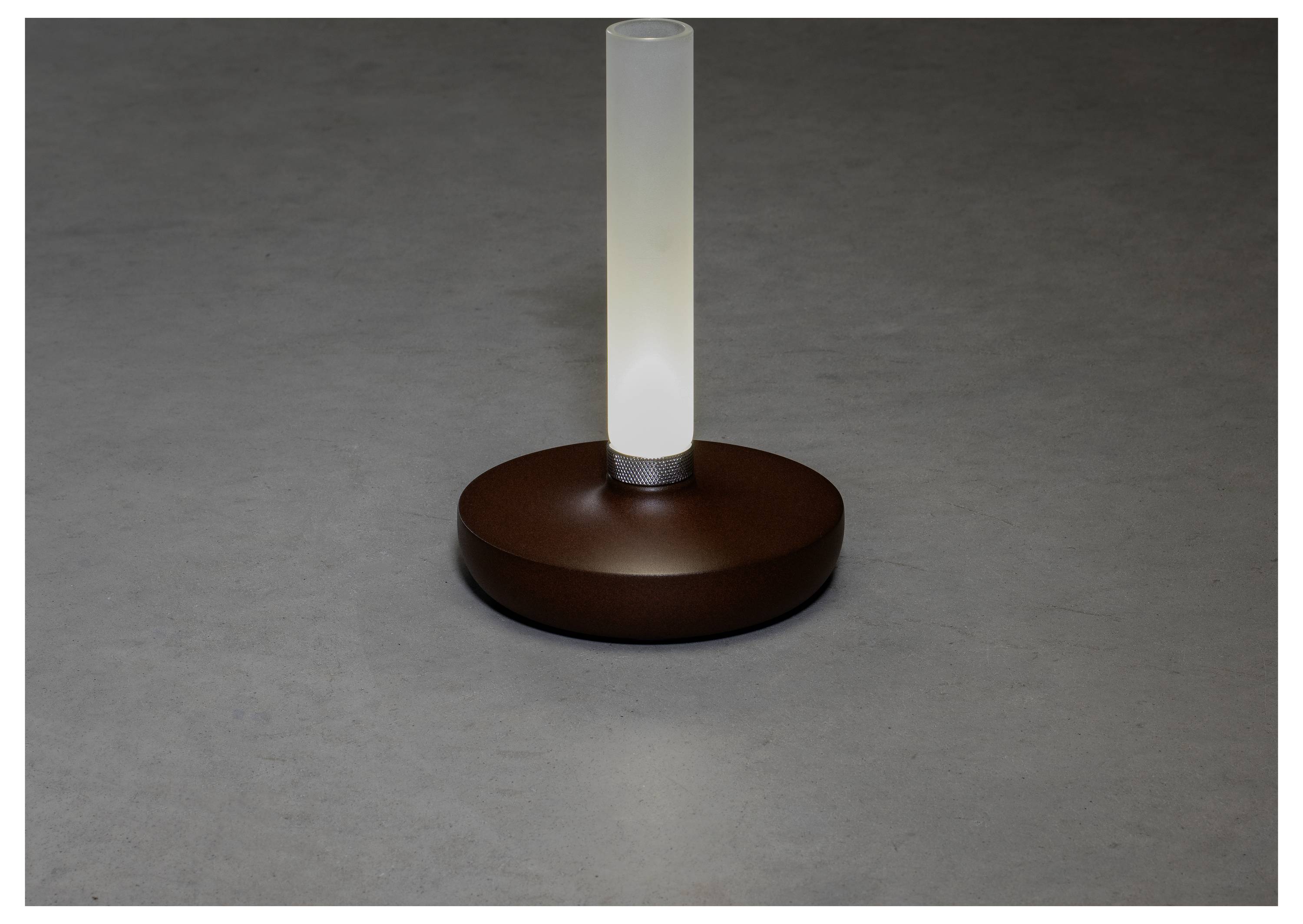 Una lampada da tavolo moderna con una lampadina cilindrica in vetro smerigliato su una base rotonda marrone, che diffonde un bagliore sottile in una stanza poco illuminata.