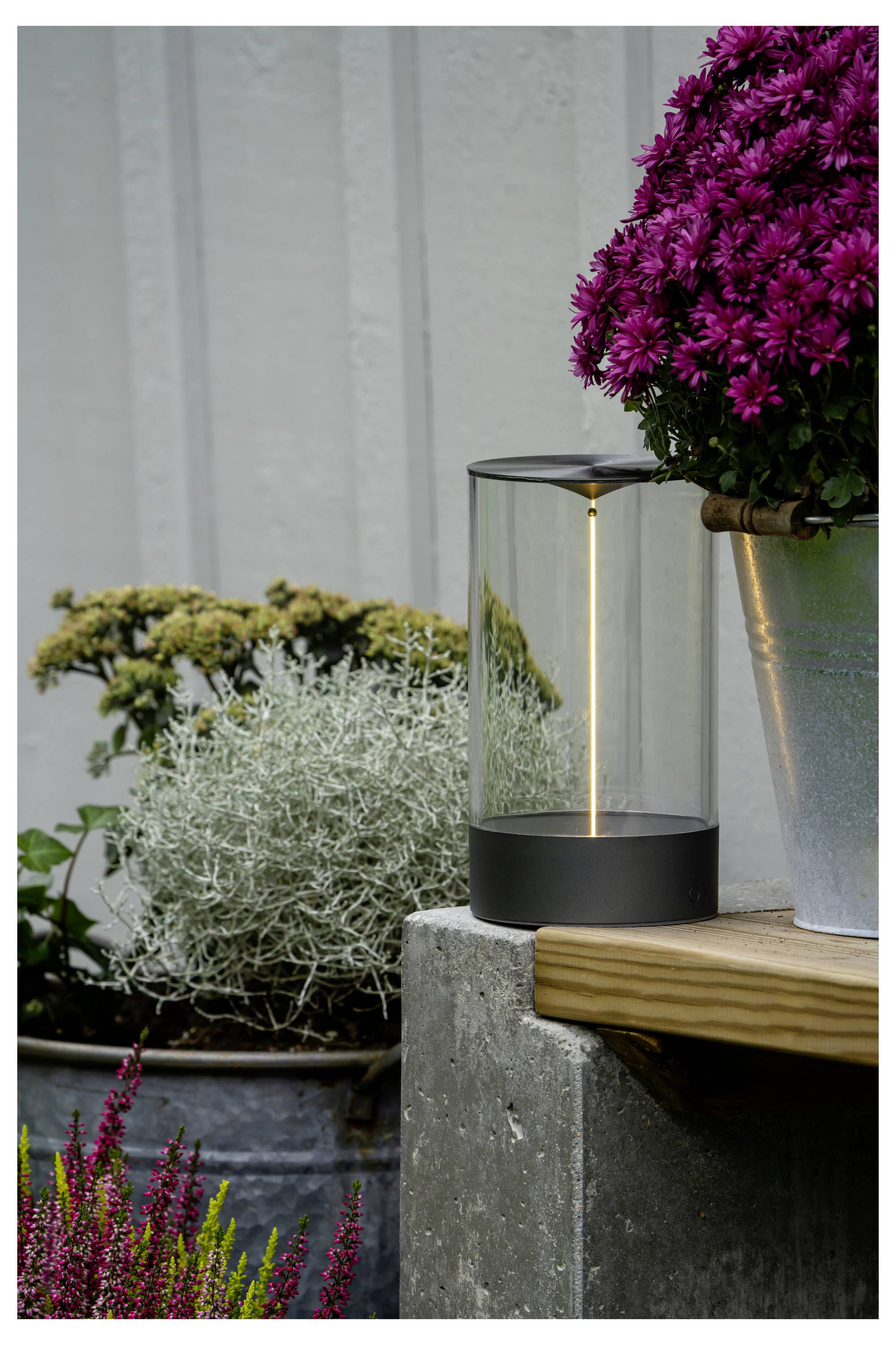 Una lampada cilindrica moderna emette una luce soffusa, posizionata vicino a fiori viola in un secchio, poggiata su un blocco di cemento all'aperto, contro un muro di legno.