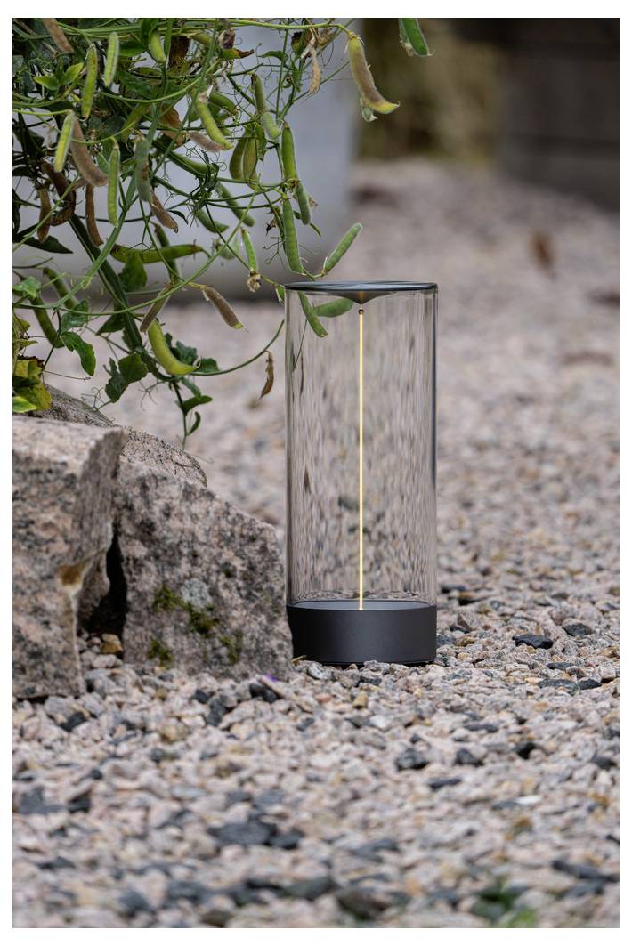 Una moderna lampada cilindrica da esterno con un design elegante, che emette una luce soffusa, è posizionata sulla ghiaia accanto alla vegetazione e a una roccia.