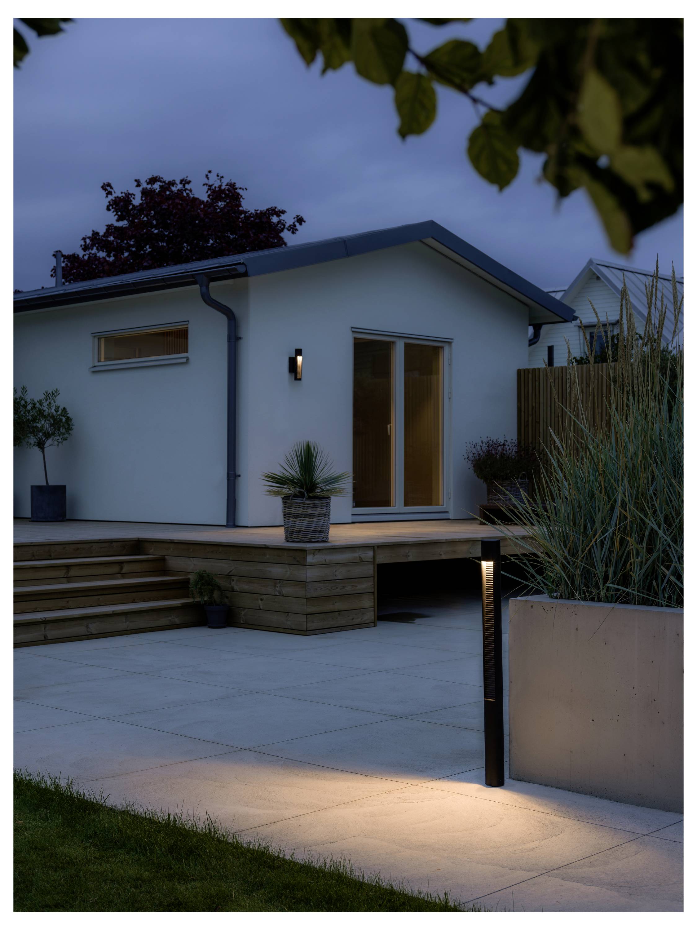 Casa moderna con un patio illuminato al crepuscolo. Un ponte in legno, piante in vaso e luci da esterno creano un'atmosfera accogliente e invitante.