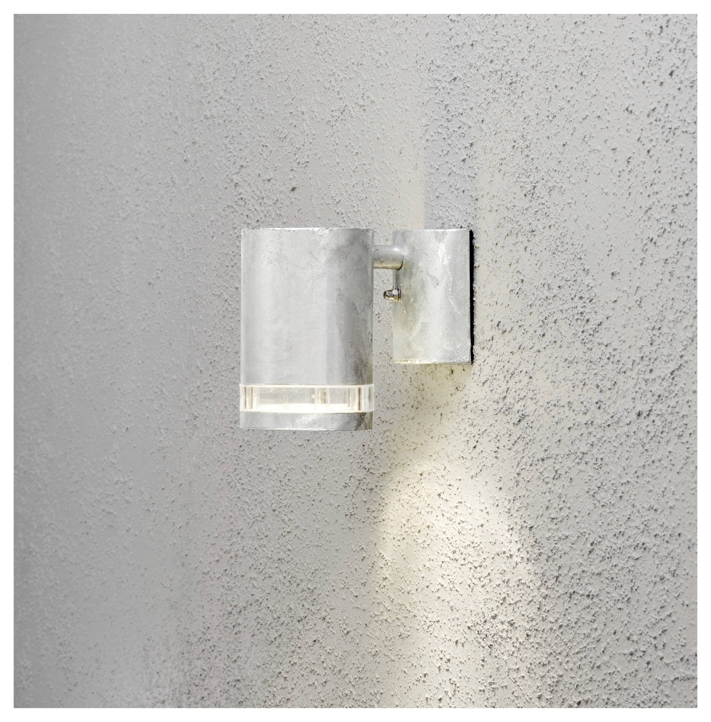 Una luce da parete esterna con un design cilindrico in metallo proietta la luce verso il basso su una superficie muraria dalla texture marcata.