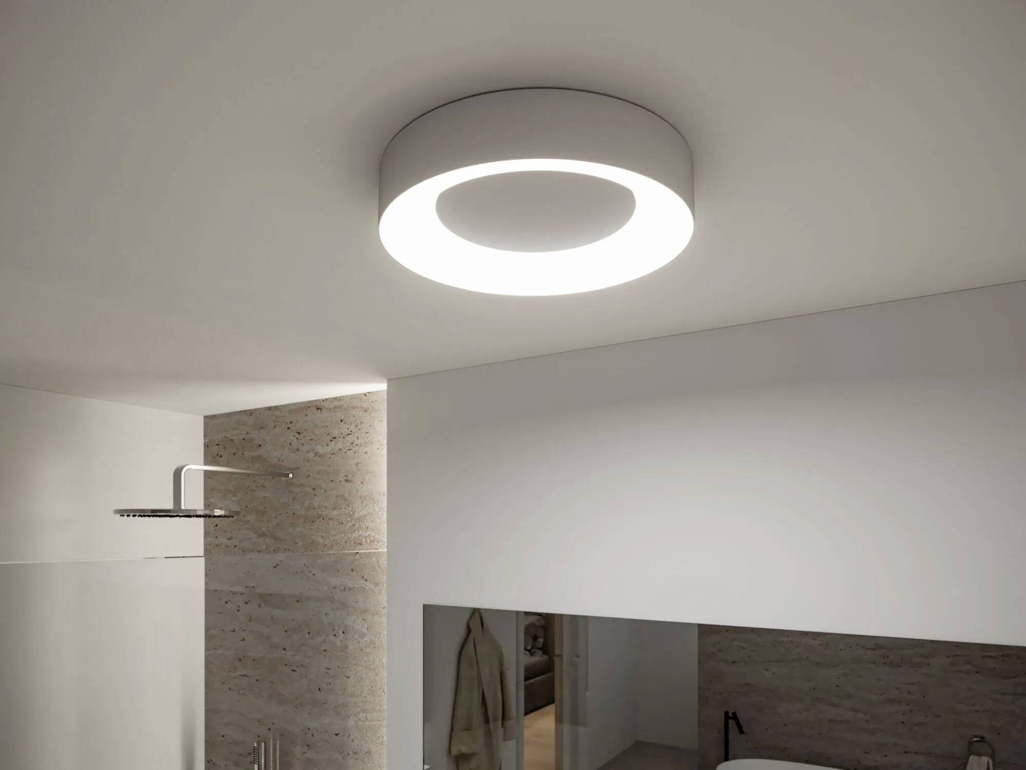 Lampada LED circolare a soffitto, illuminata in un bagno moderno con pareti piastrellate beige e soffione della doccia visibile.