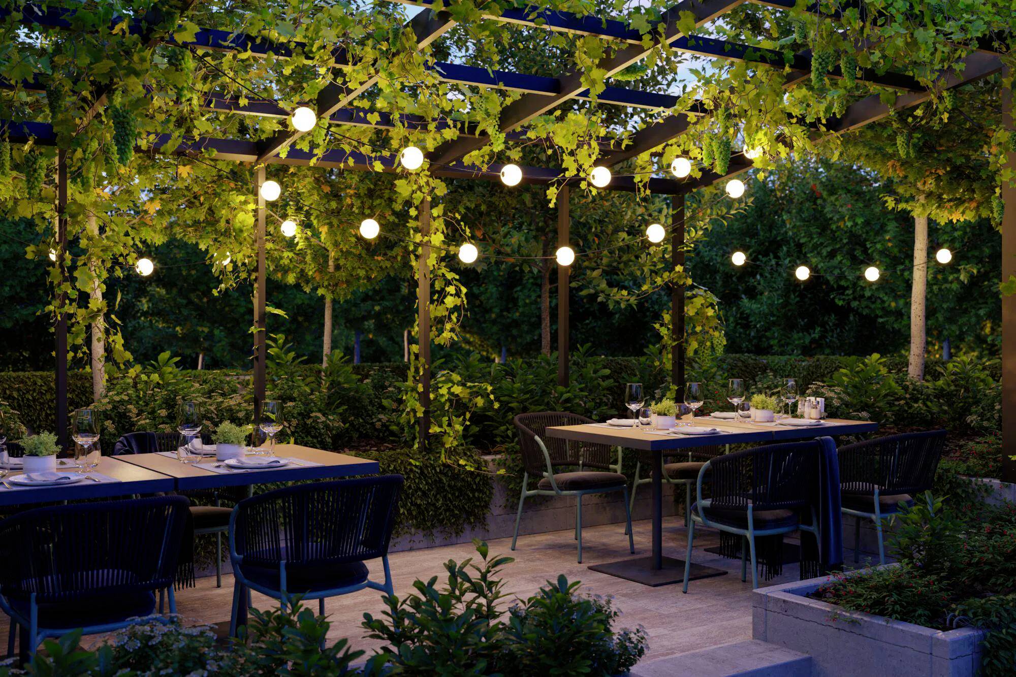 Area ristorante all'aperto con tavoli e sedie sotto una pergola decorata con luci a festone e rigogliosa vegetazione, che crea un'atmosfera accogliente.
