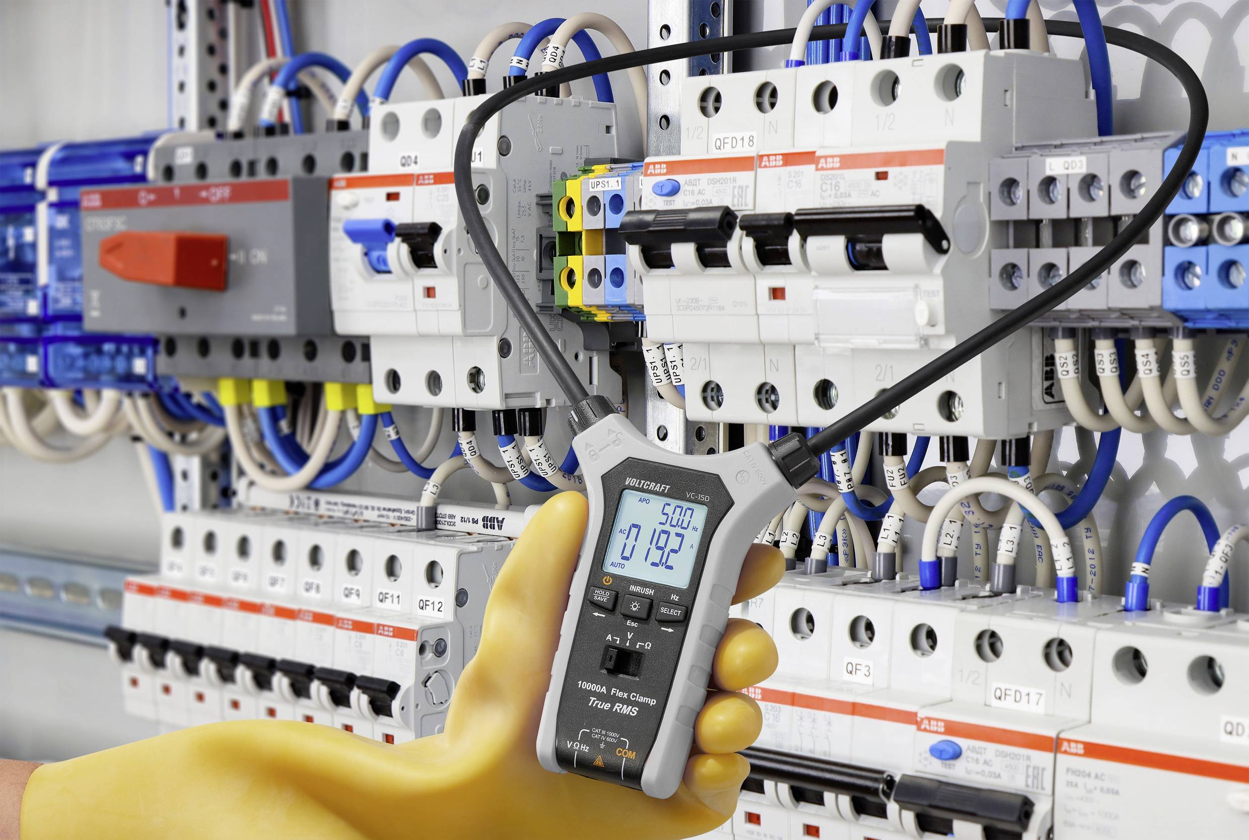 Una persona che indossa guanti isolanti utilizza un multimetro digitale per misurare la tensione su un grande quadro elettrico riempito di fili e interruttori.