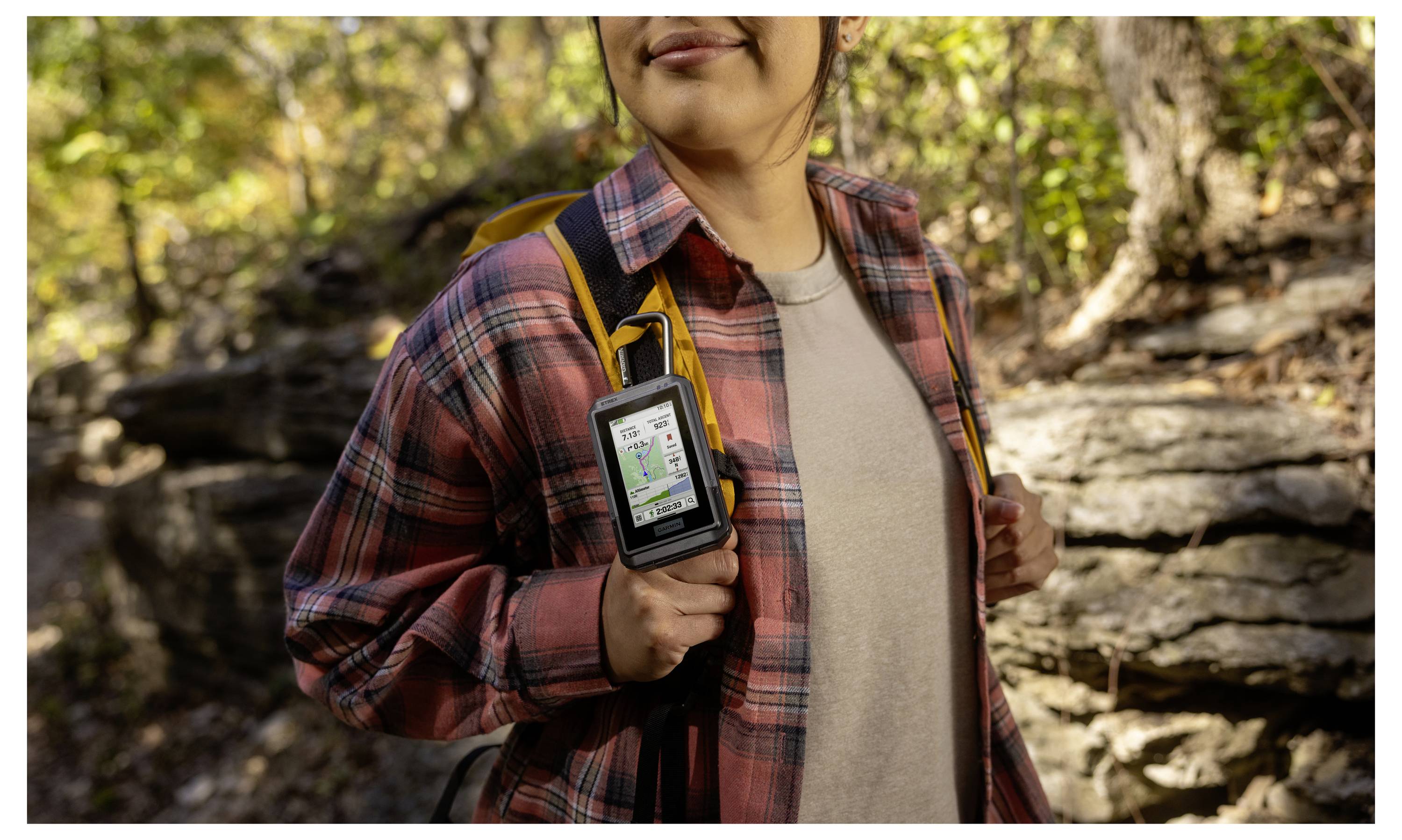 Una persona che fa escursionismo in un bosco utilizza un dispositivo GPS portatile che mostra una mappa, indossa una camicia a quadri e uno zaino, circondato da alberi.