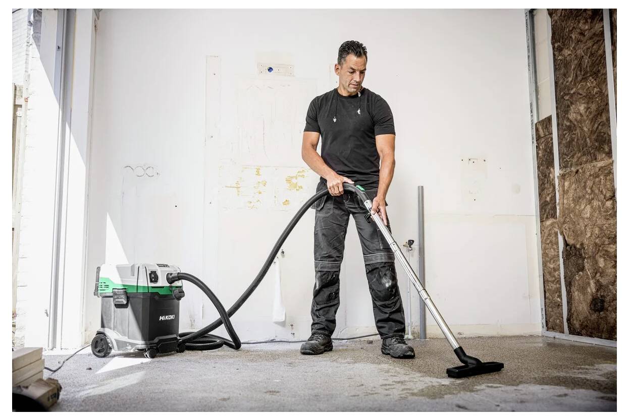 Una persona aspira un pavimento in una stanza parzialmente ristrutturata utilizzando un aspirapolvere industriale, indossando un abbigliamento da lavoro casual.