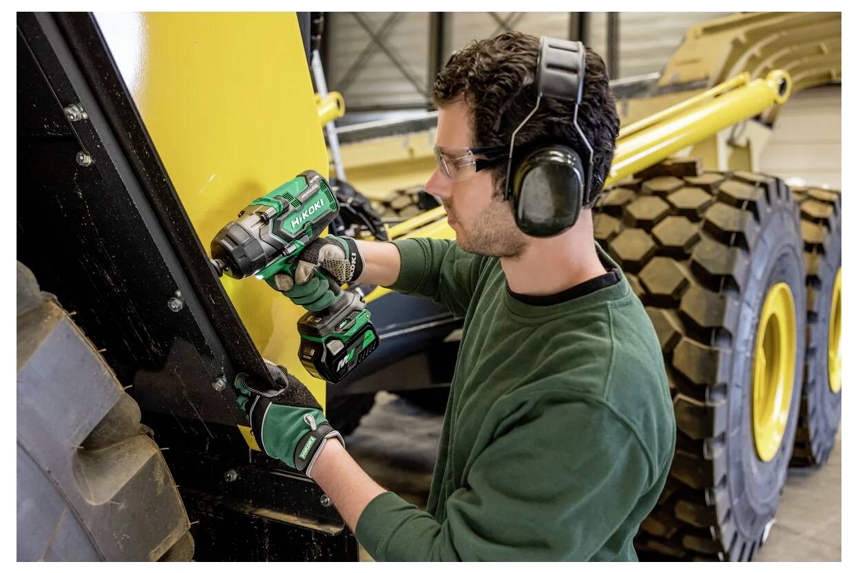 Un uomo indossa un equipaggiamento di sicurezza mentre usa un trapano elettrico su un macchinario. È concentrato sull'attività, con grandi parti di veicoli visibili sullo sfondo.