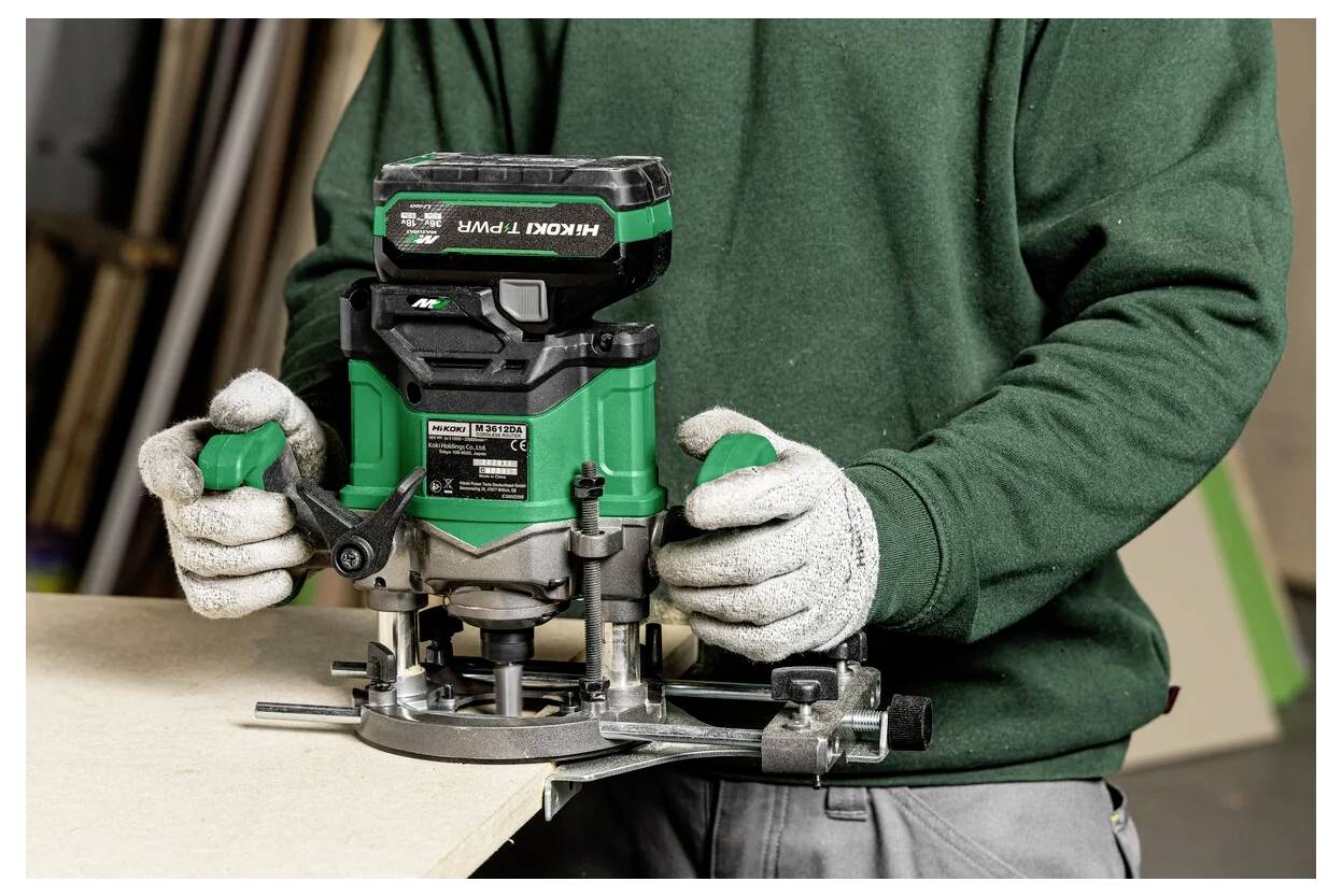 Una persona con i guanti utilizza un router verde su una tavola di legno, concentrandosi su un lavoro di artigianato o falegnameria in un ambiente di officina.