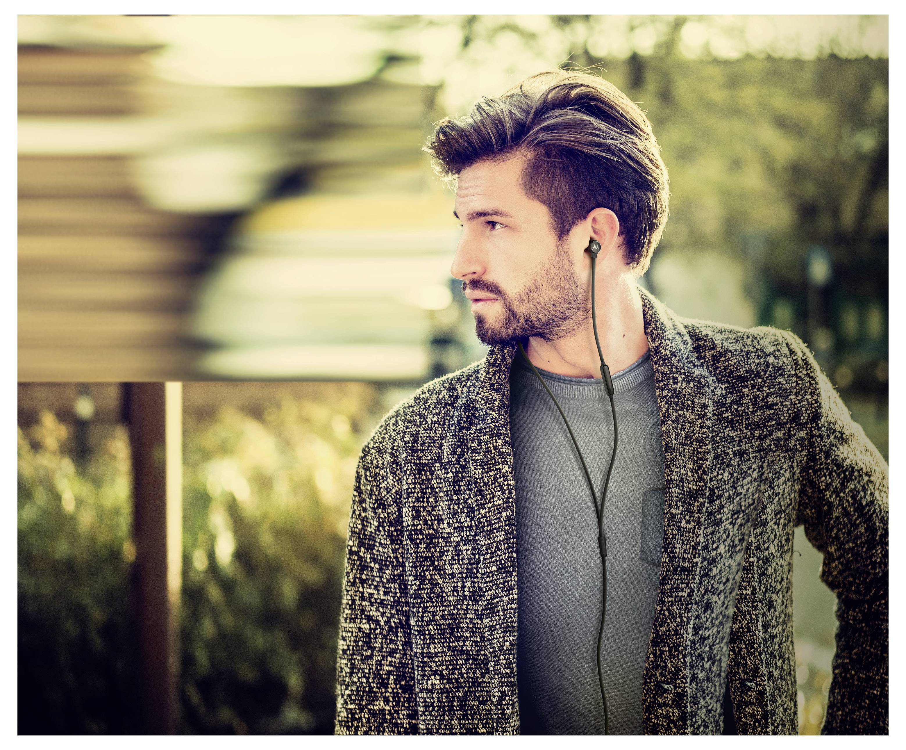 Un uomo con la barba e gli auricolari guarda di lato all'aperto. La sua espressione è pensierosa. La vegetazione sullo sfondo è sfocata.