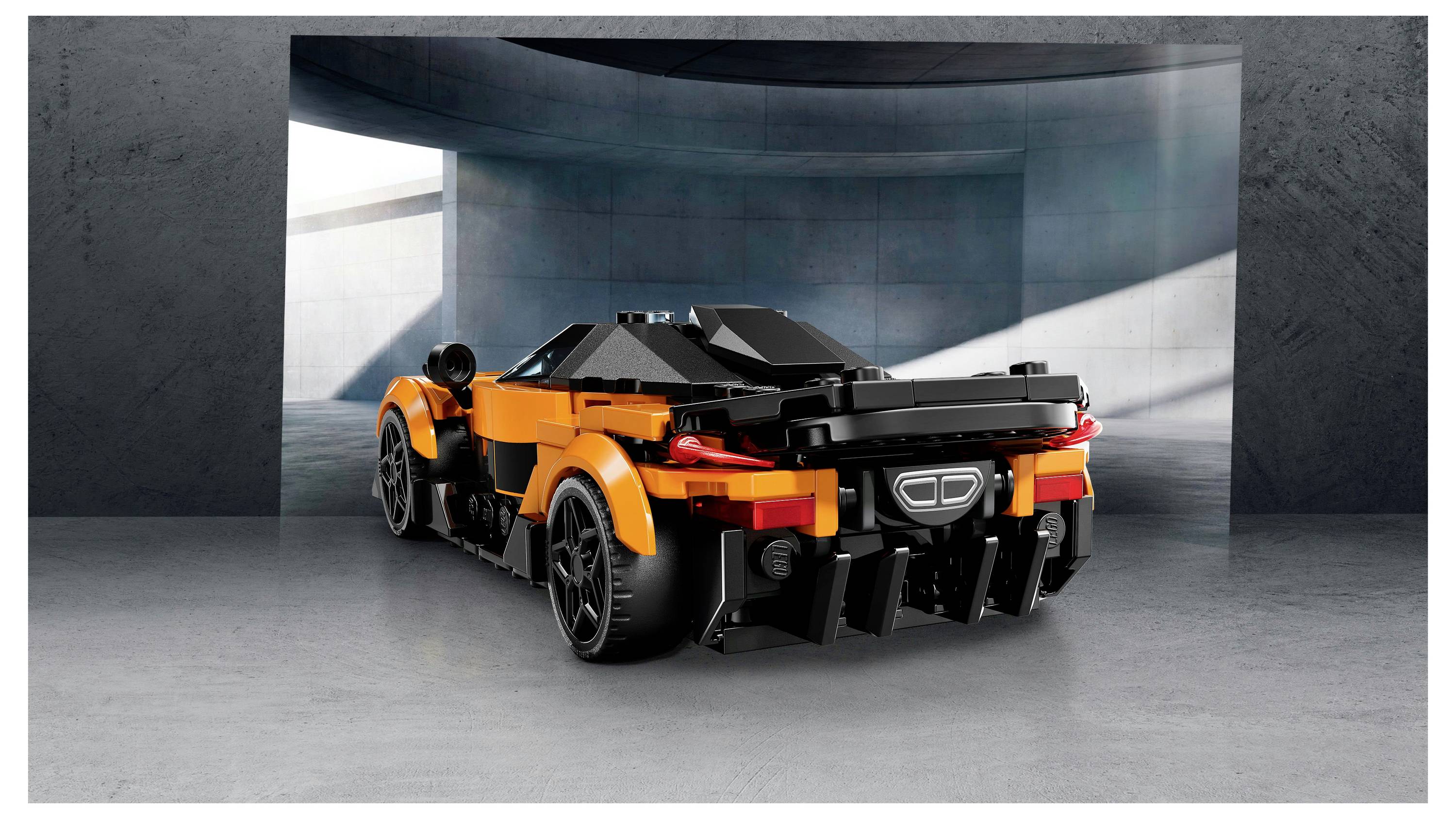 Un modello di auto sportiva arancione e nera, costruito con mattoncini interconnessi, è esposto in un ambiente garage di cemento, illuminato da una morbida luce diurna.