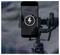 Un smartphone montato su un gimbal mostra un'icona di ricarica sopra un'immagine paesaggistica con una figura distante su una collina.