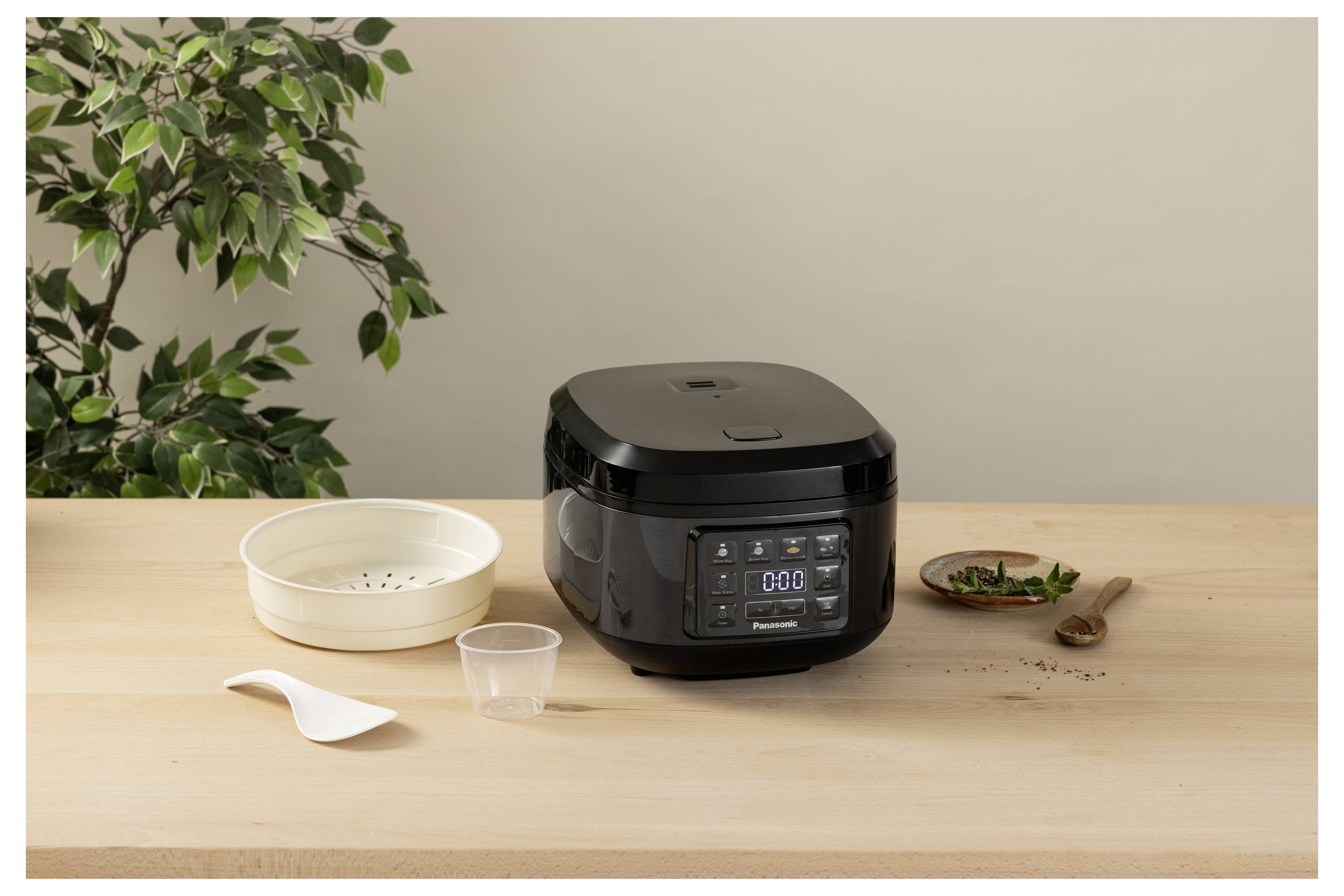 Un multicooker nero su un piano di lavoro in legno con accessori da cucina. Una pianta sullo sfondo crea un ambiente domestico e accogliente.