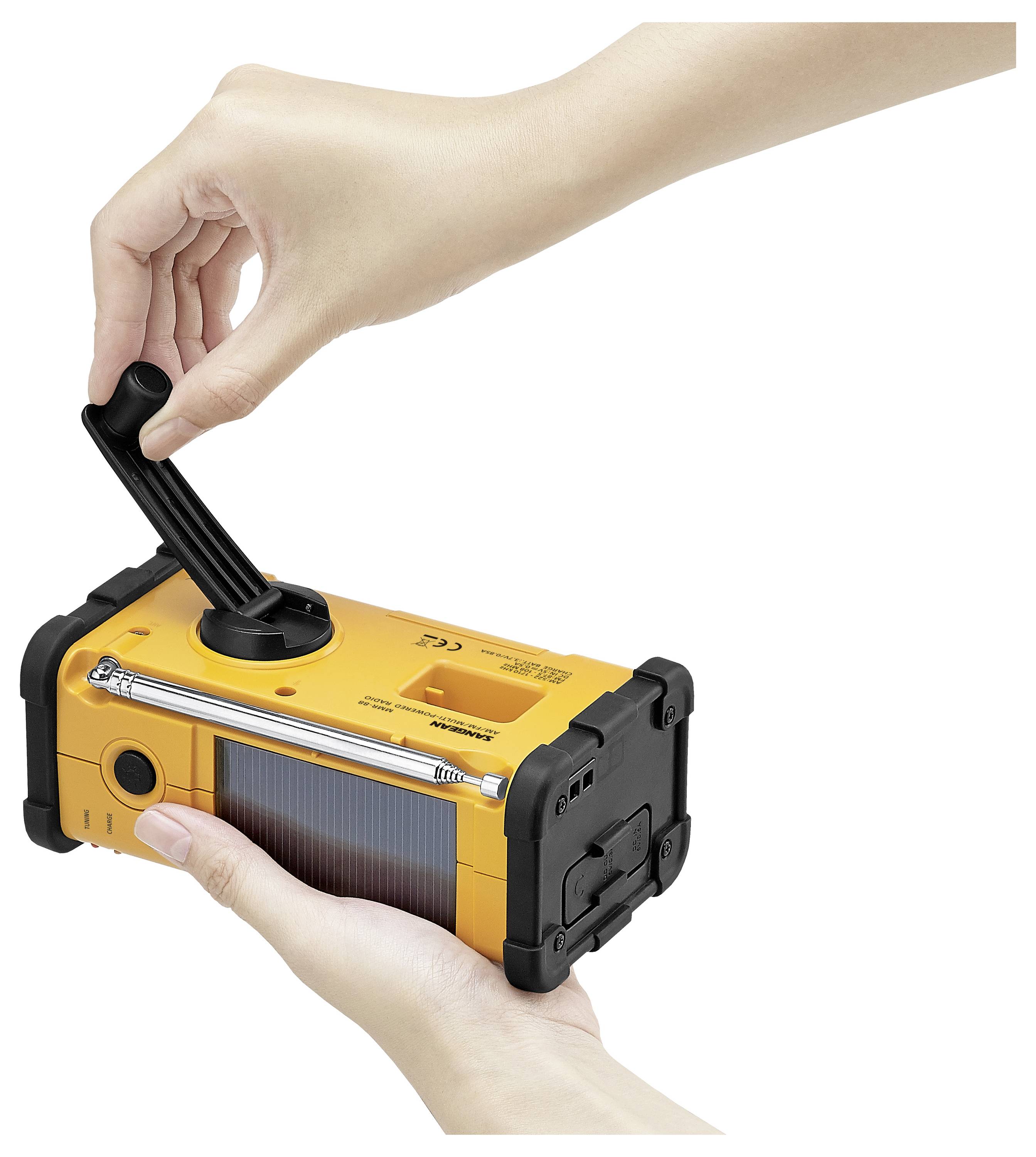 Primo piano di mani che utilizzano una radio di emergenza gialla con manovella e pannello solare, dimostrando la sua funzione di ricarica manuale.