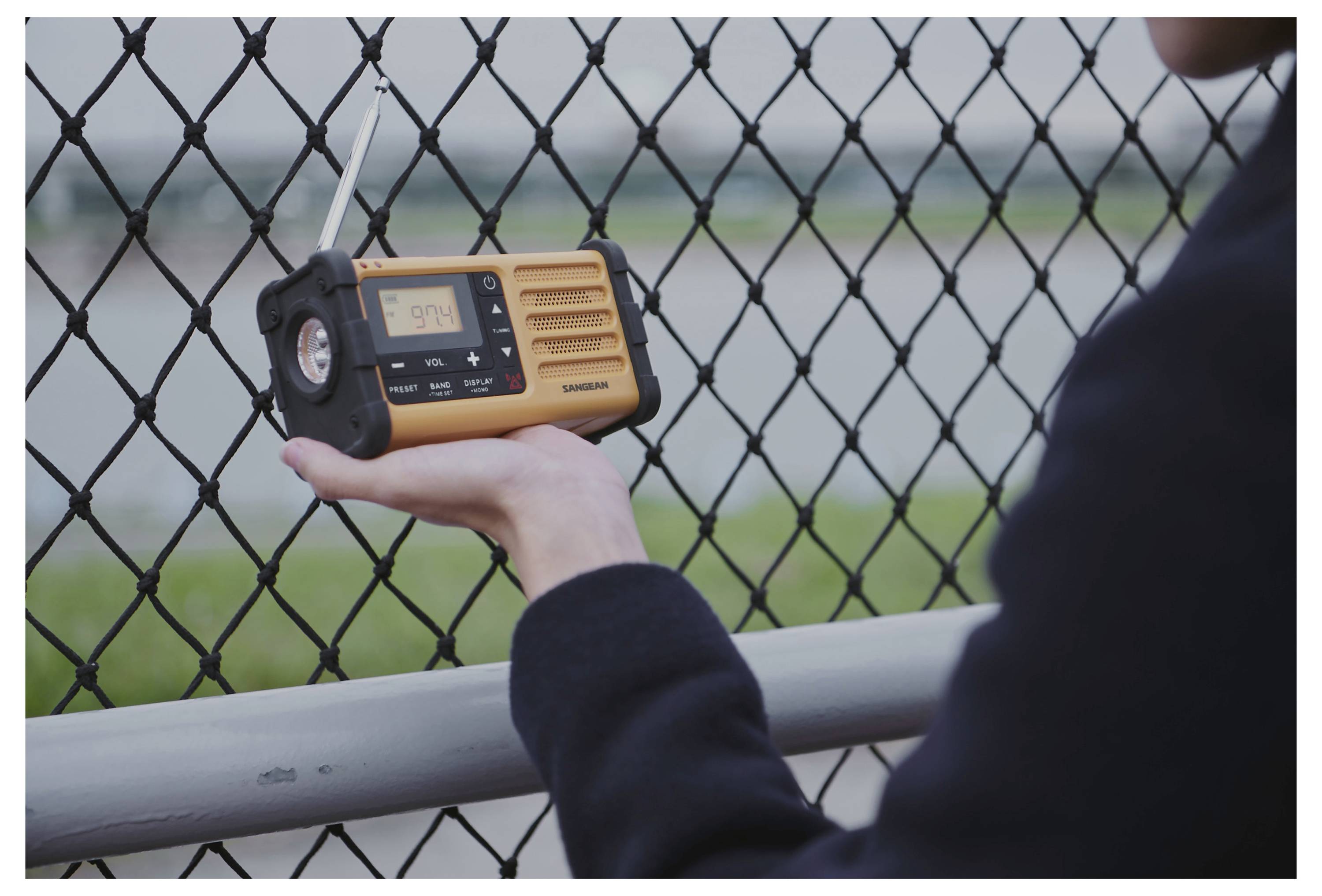 Una persona tiene in mano una radio di emergenza gialla con un display che mostra '5:14'. Sullo sfondo c'è una recinzione e un paesaggio sfocato.
