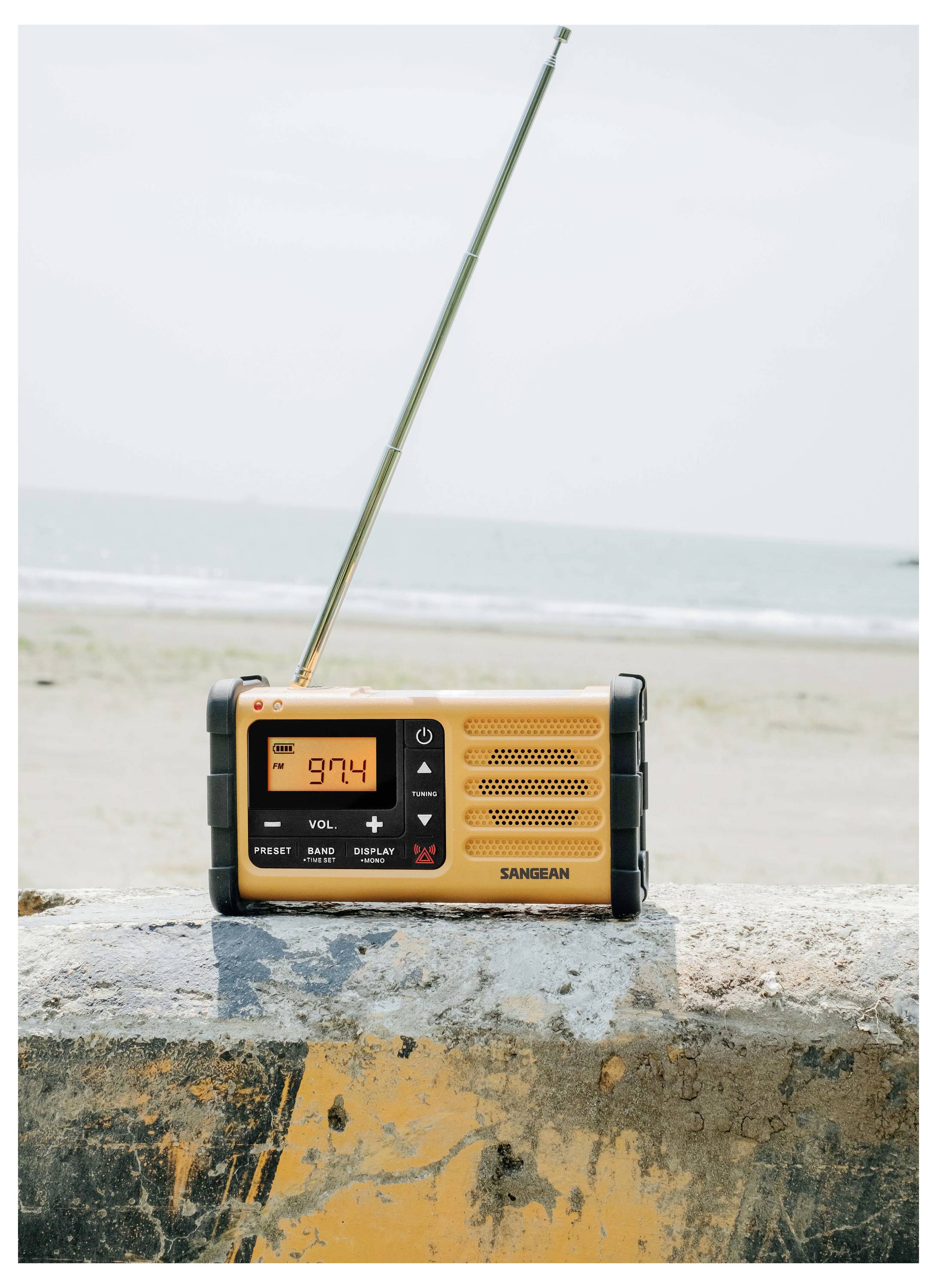 Una radio portatile gialla con antenna estesa è poggiata su una barriera di cemento vicino alla spiaggia. Il display mostra '97.4'.
