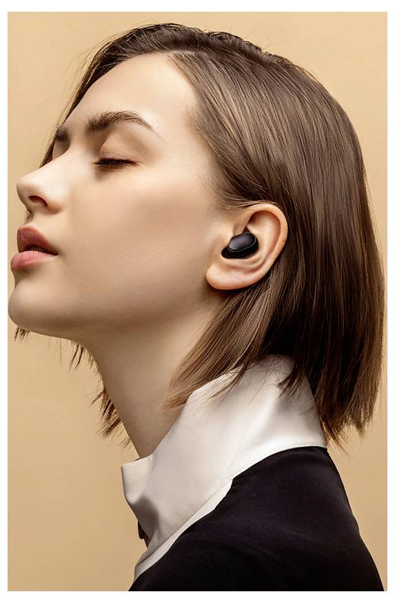 Una persona con capelli corti marroni indossa auricolari wireless neri. Ha un'espressione calma e lo sfondo è di un caldo colore beige.