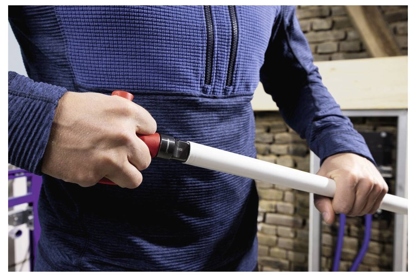 Una persona con un maglione blu sta tagliando un tubo bianco con un tagliatubo rosso sullo sfondo di pietre impilate e travi di legno.