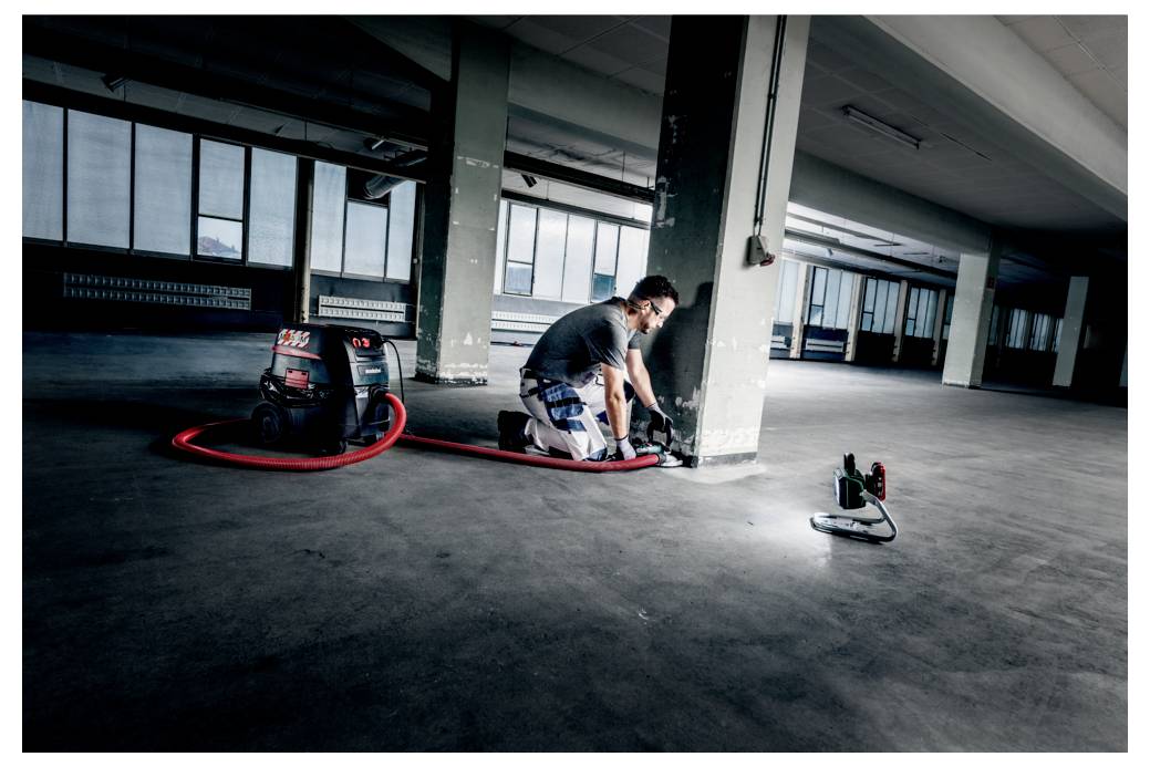 Una persona sta riparando un muro in un edificio spazioso e vuoto, con un tubo dell'aspirapolvere rosso e una lampada da lavoro sul pavimento.