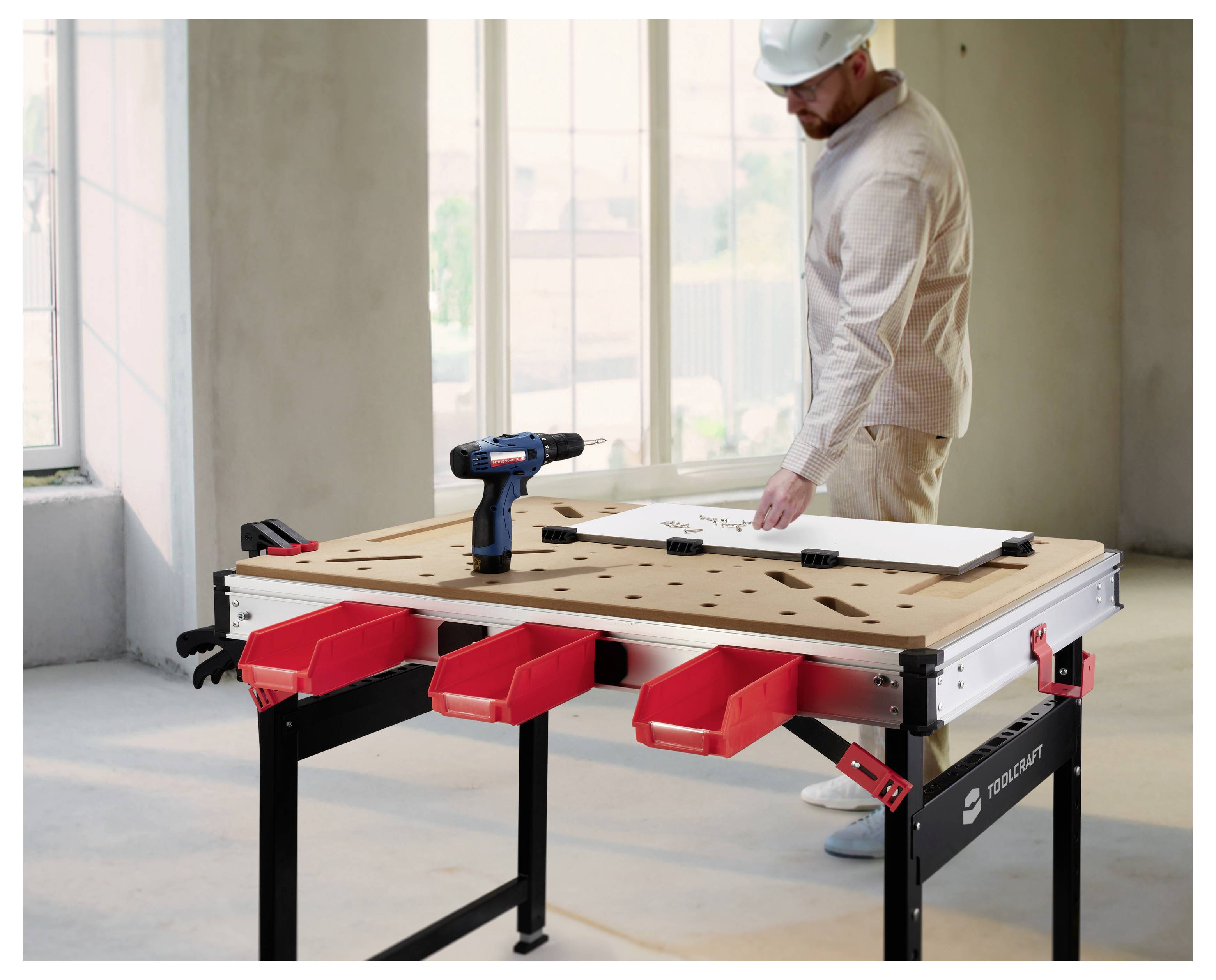 Operaio edile con casco di protezione che misura un pannello bianco su un banco da lavoro con un trapano elettrico e contenitori di stoccaggio rossi in una stanza non ancora completata.