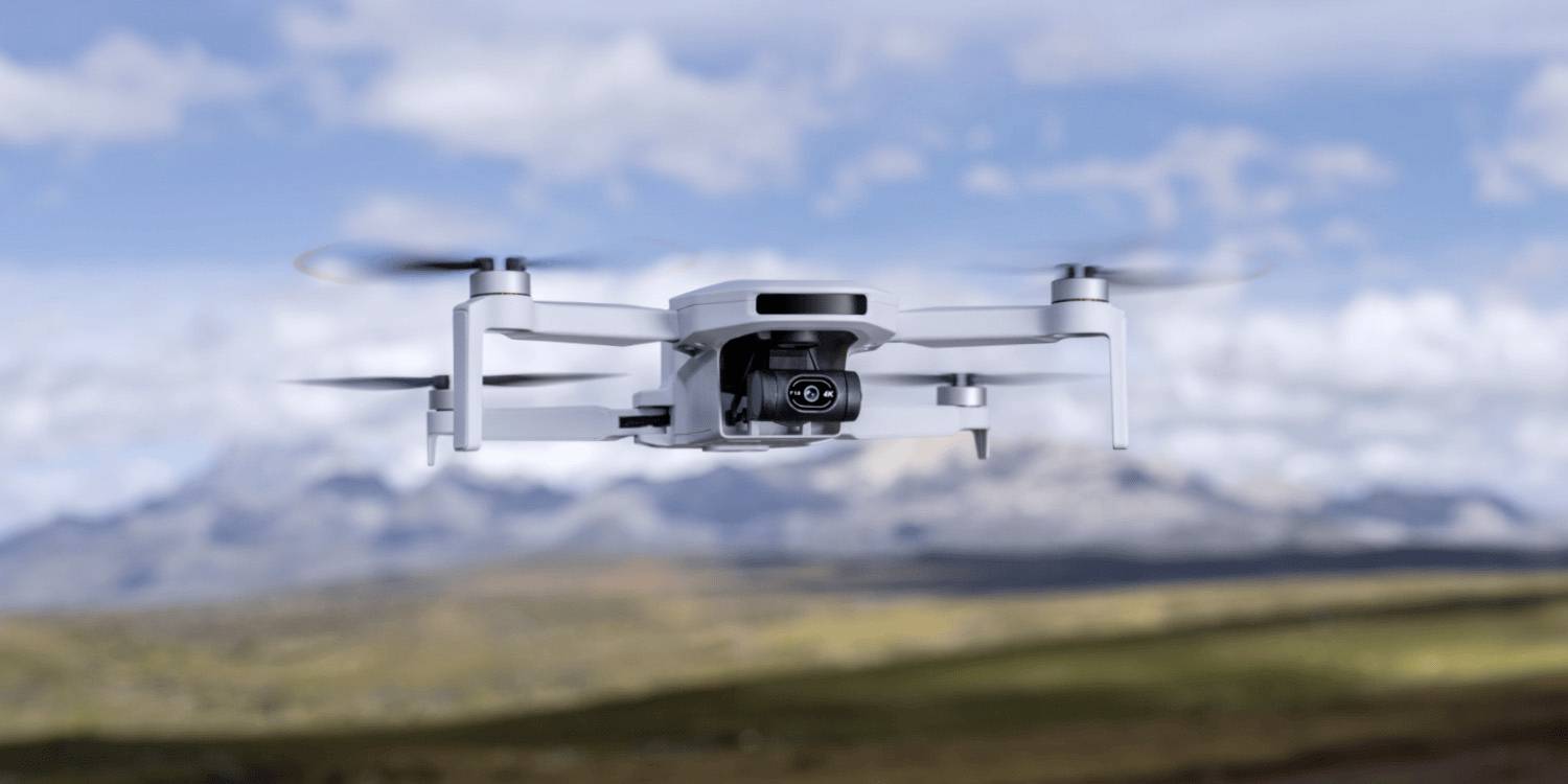 Un drone bianco con una fotocamera aleggia in primo piano, con un paesaggio montano sfocato e un cielo blu sullo sfondo.
