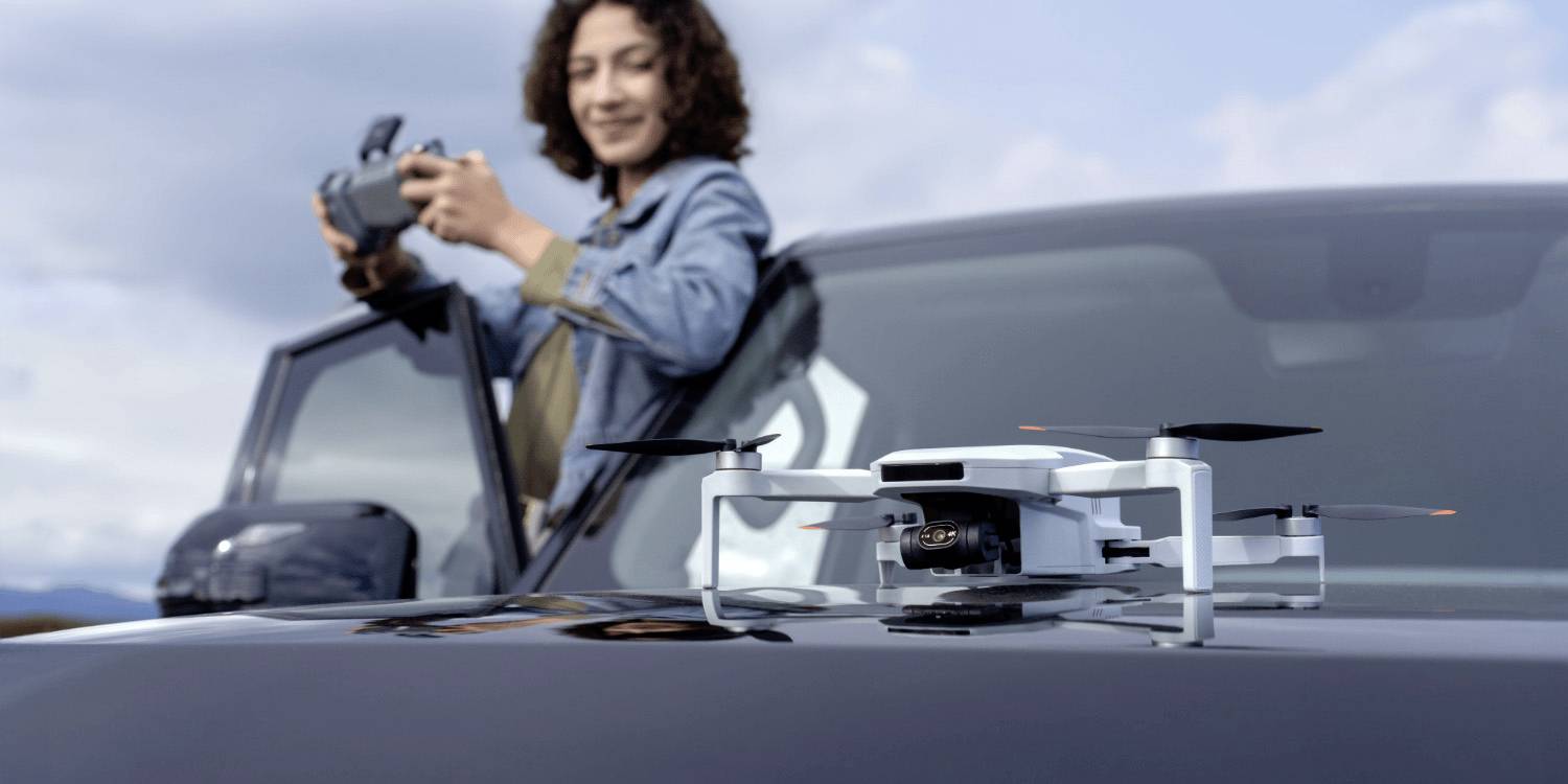 Una persona controlla un drone con un telecomando, in piedi accanto a un SUV. Il drone è sul cofano del veicolo, pronto per decollare.