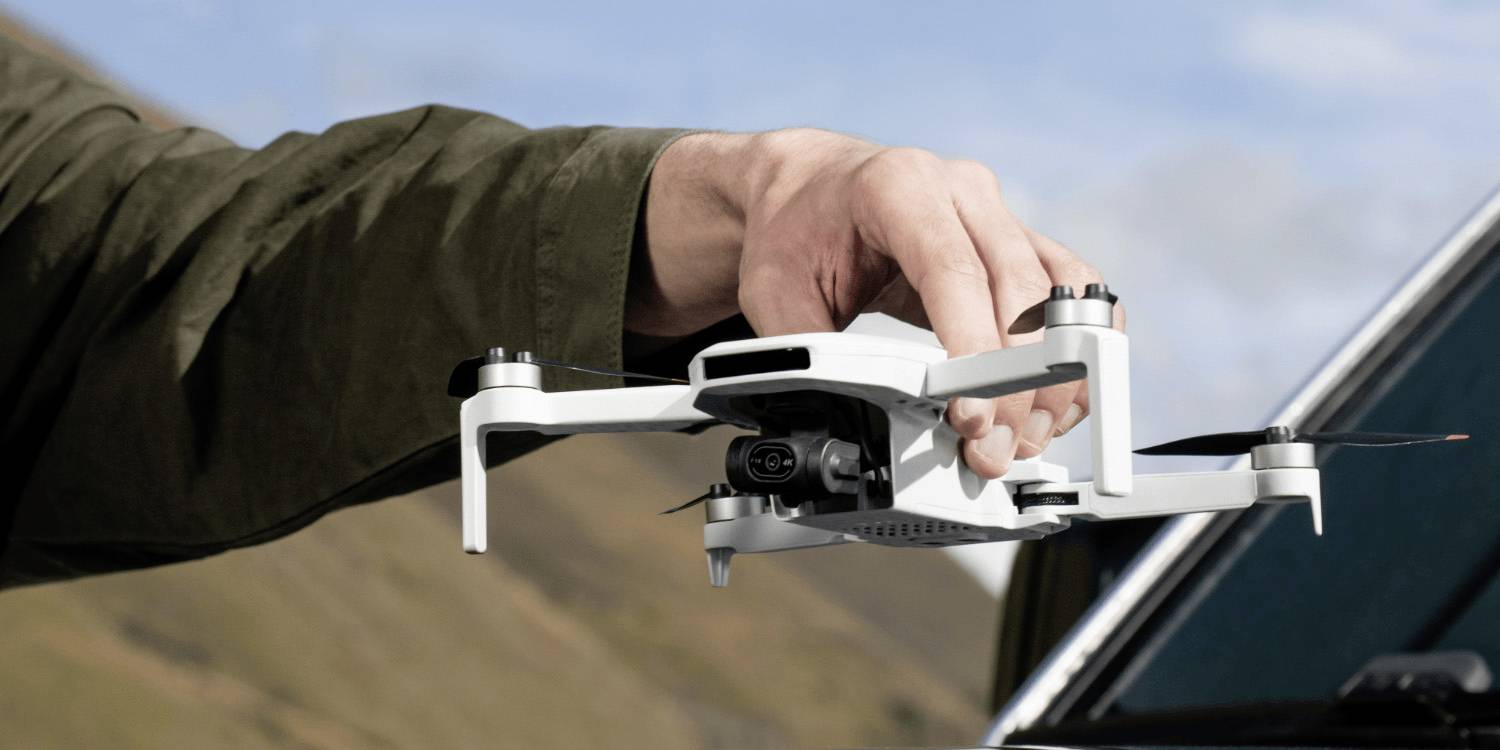 Una persona con una giacca verde tiene in mano un piccolo drone in un ambiente esterno, suggerendo la preparazione per un volo. Sullo sfondo si vedono colline sotto un cielo blu.