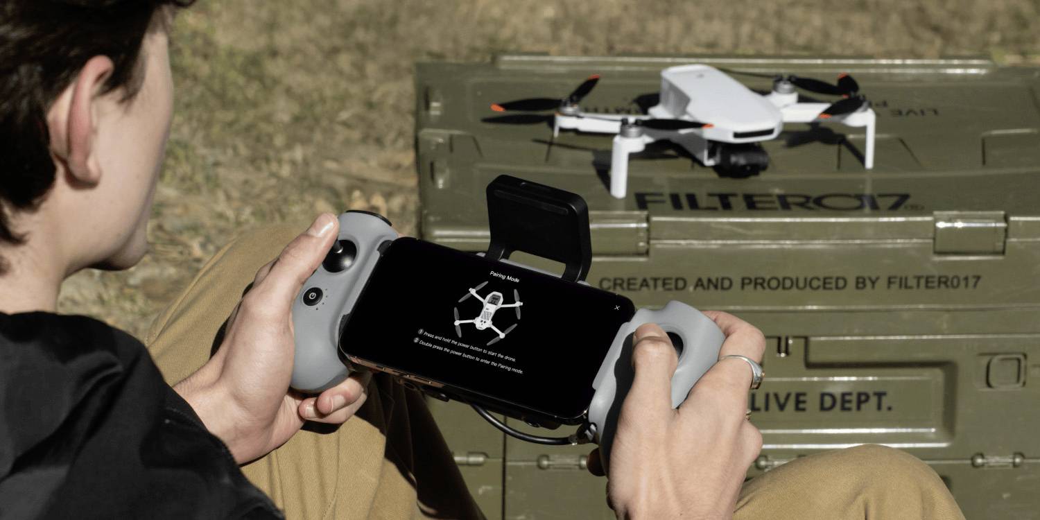 Una persona manovra un drone utilizzando un controller portatile e uno smartphone. Il drone è posizionato su un contenitore metallico verde etichettato 'FILTER017.'