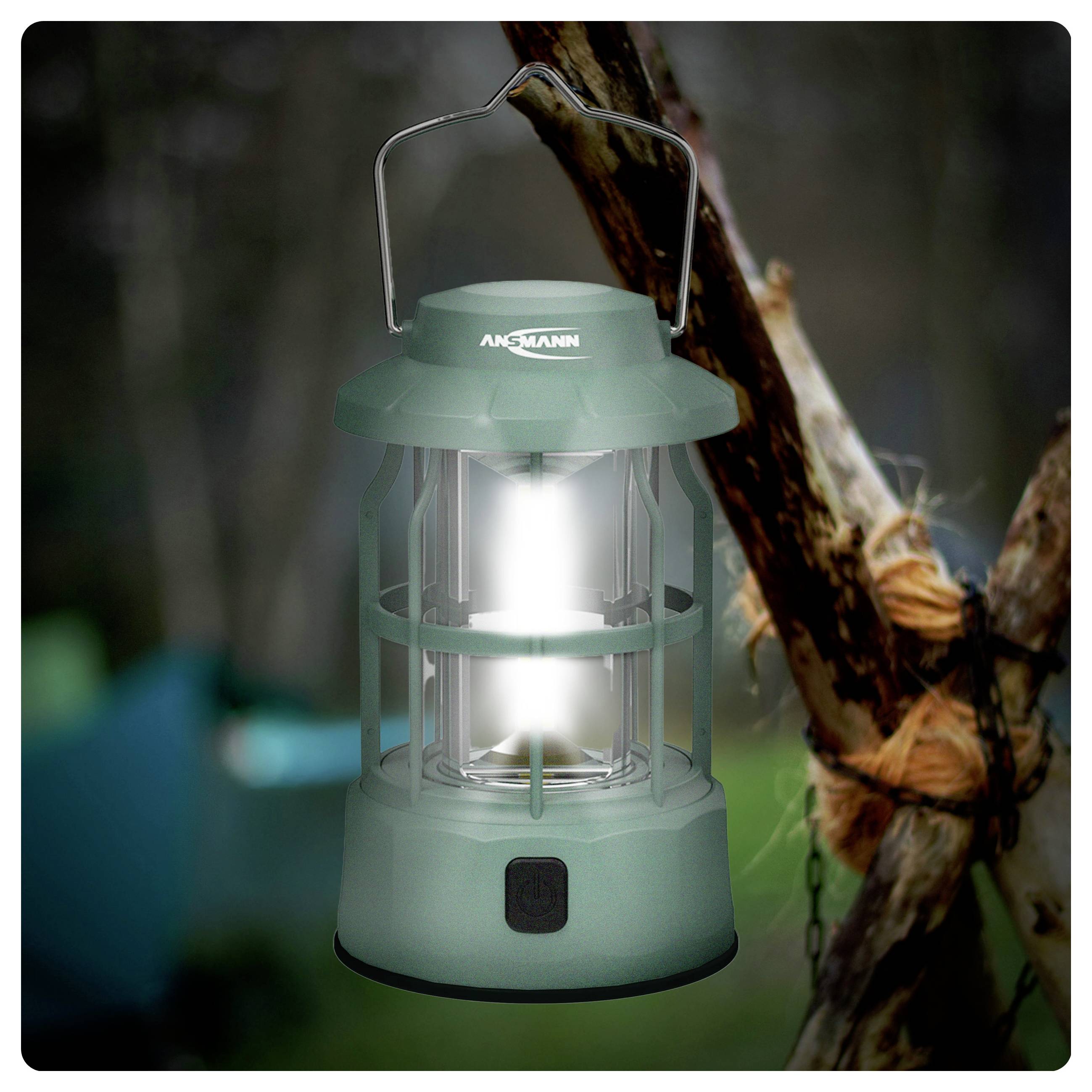 Una lanterna da campeggio verde appesa a un ramo di un albero, illuminata luminosamente. L'area circostante appare boschiva, indicando un ambiente esterno.