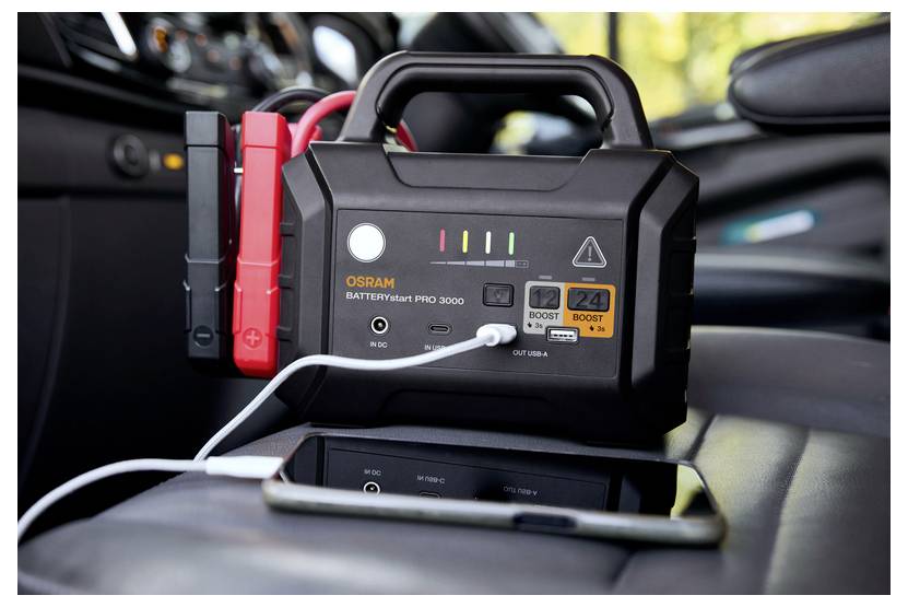 'Un avviatore di emergenza portatile con cavi collegati, poggiato su un sedile dell'auto. Uno smartphone si sta caricando tramite una porta USB del dispositivo.'