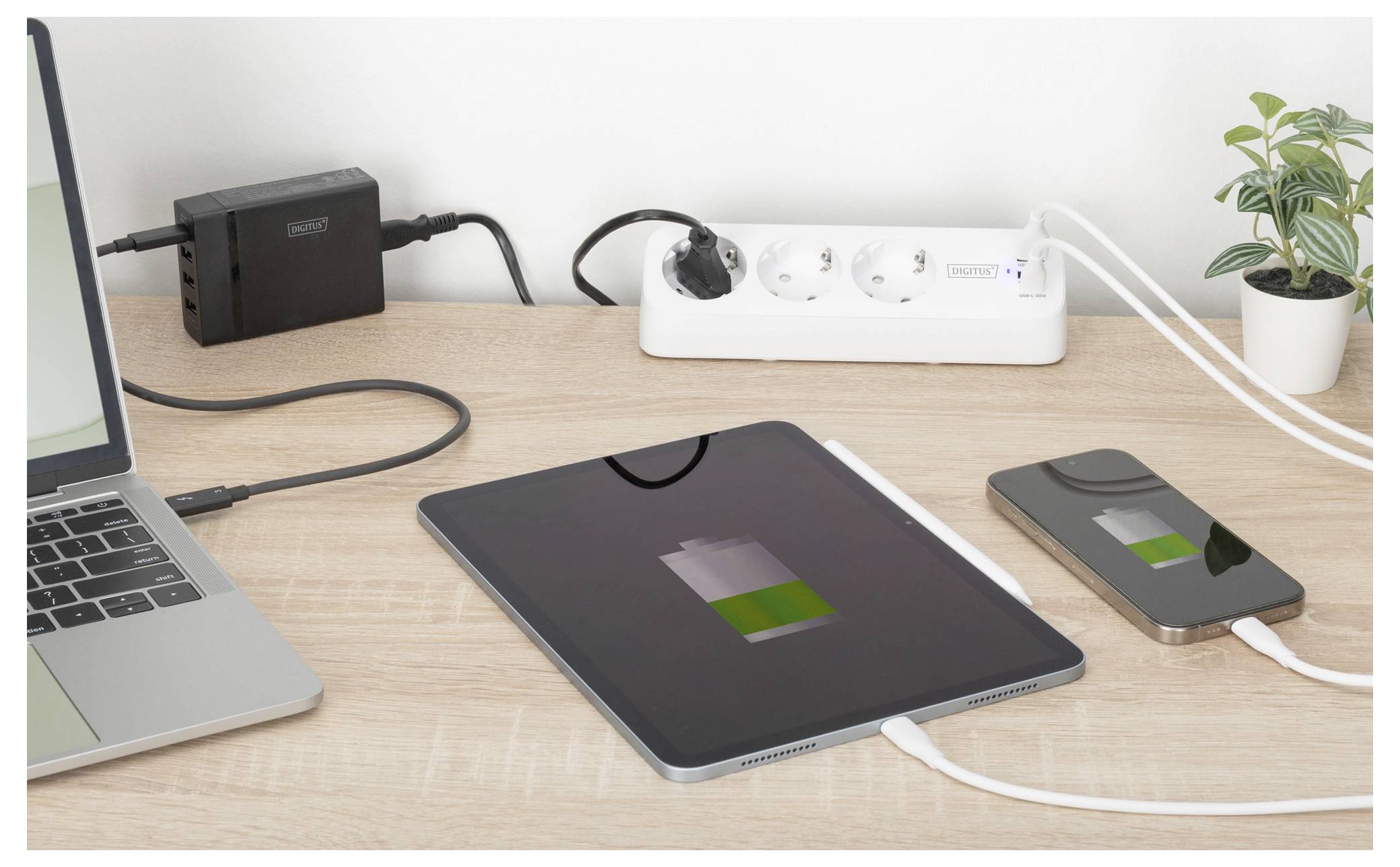 Una scrivania mostra un tablet in carica e uno smartphone collegati a una ciabatta elettrica, con una pianta e un laptop nelle vicinanze, indicando la ricarica dei dispositivi.