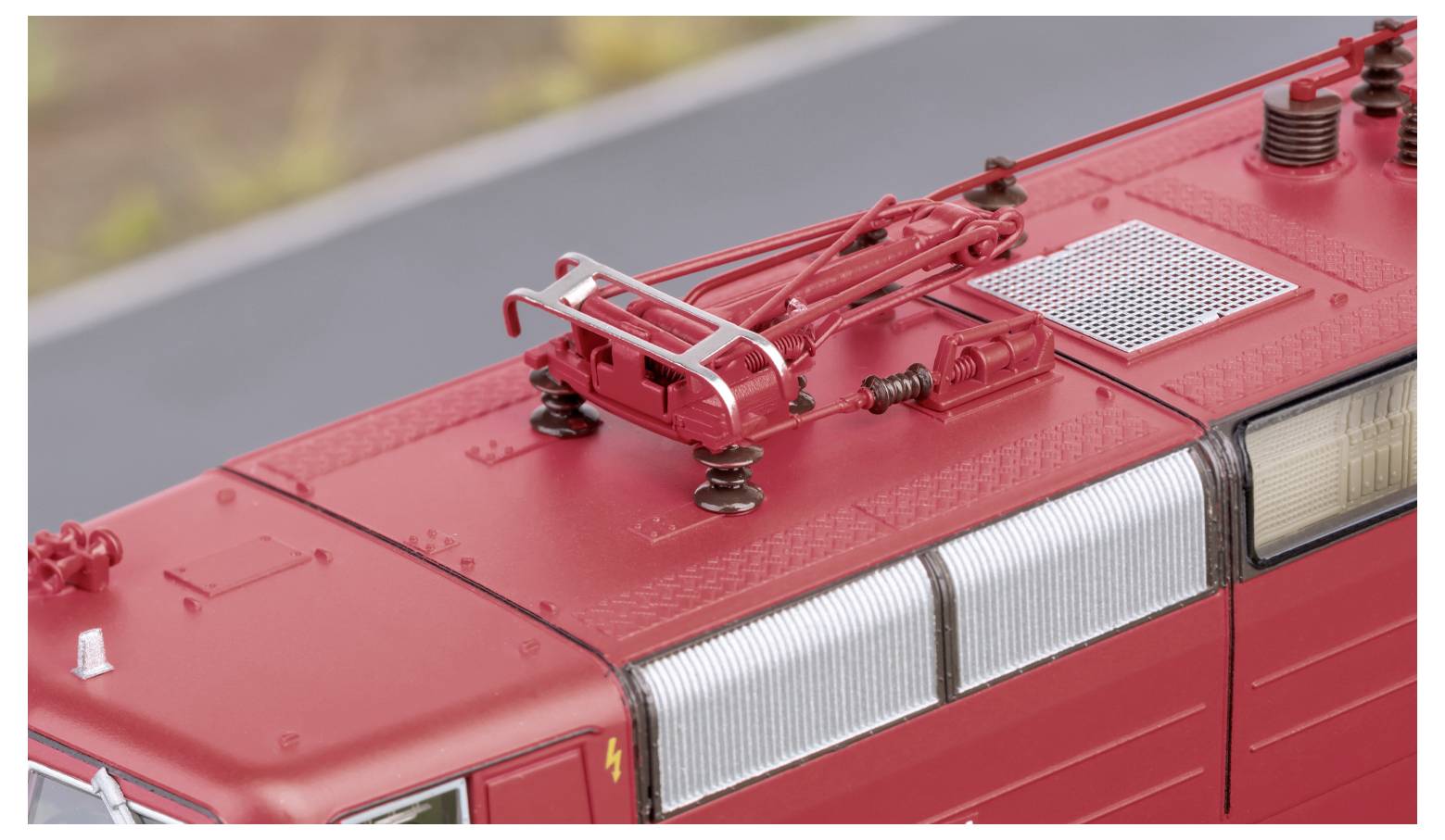 L'immagine mostra il tetto di una locomotiva elettrica rossa con pantografo e cablaggi visibili sullo sfondo sfocato.