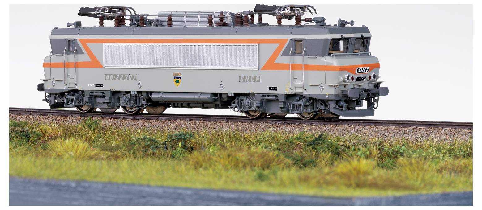 Un locomotore di un modello di treno con dettagli arancioni e grigi viaggia su un binario immerso in un paesaggio erboso.