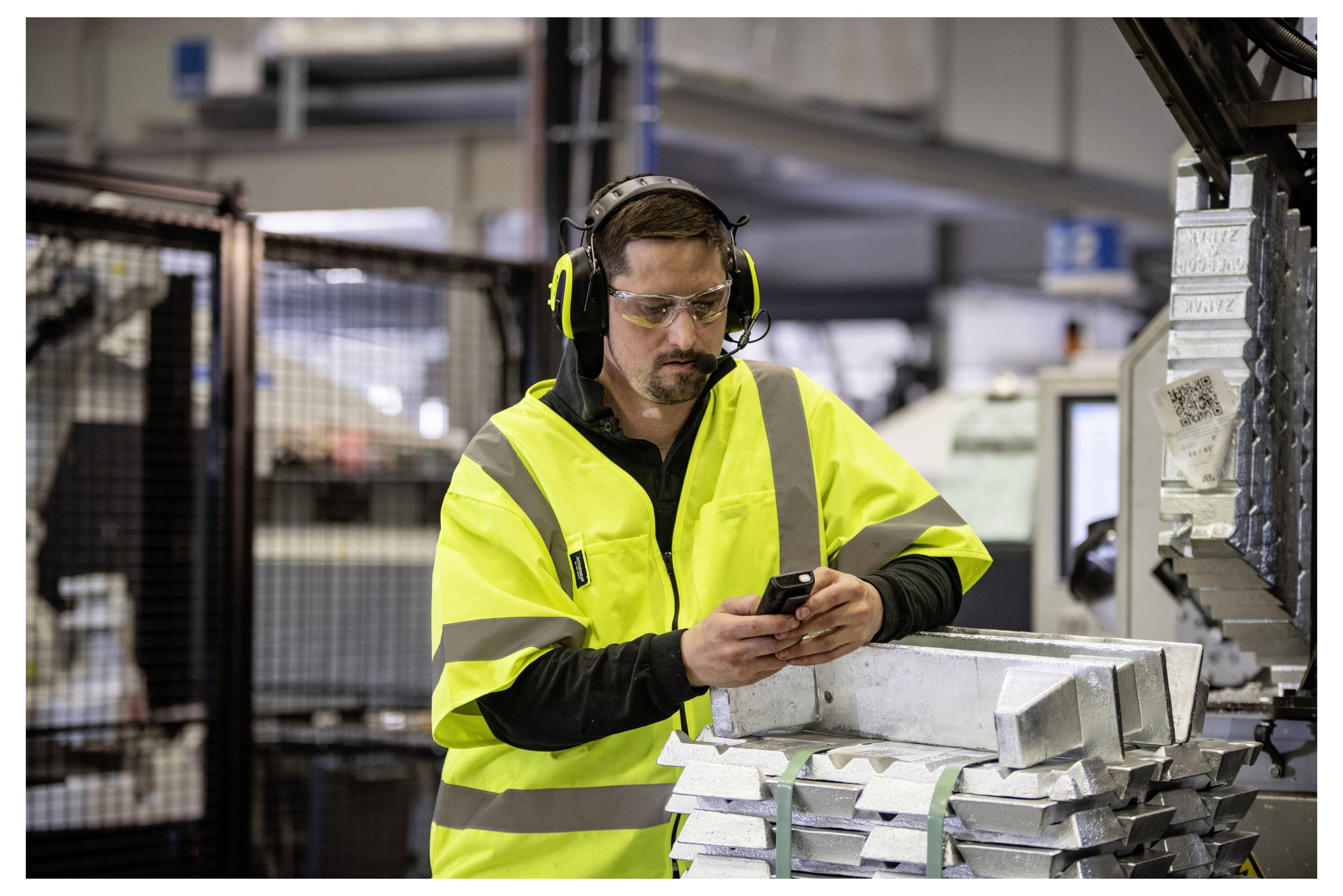 Un lavoratore con un giubbotto ad alta visibilità e dispositivi di sicurezza controlla il suo telefono mentre è in piedi accanto a materiali metallici in un ambiente industriale.
