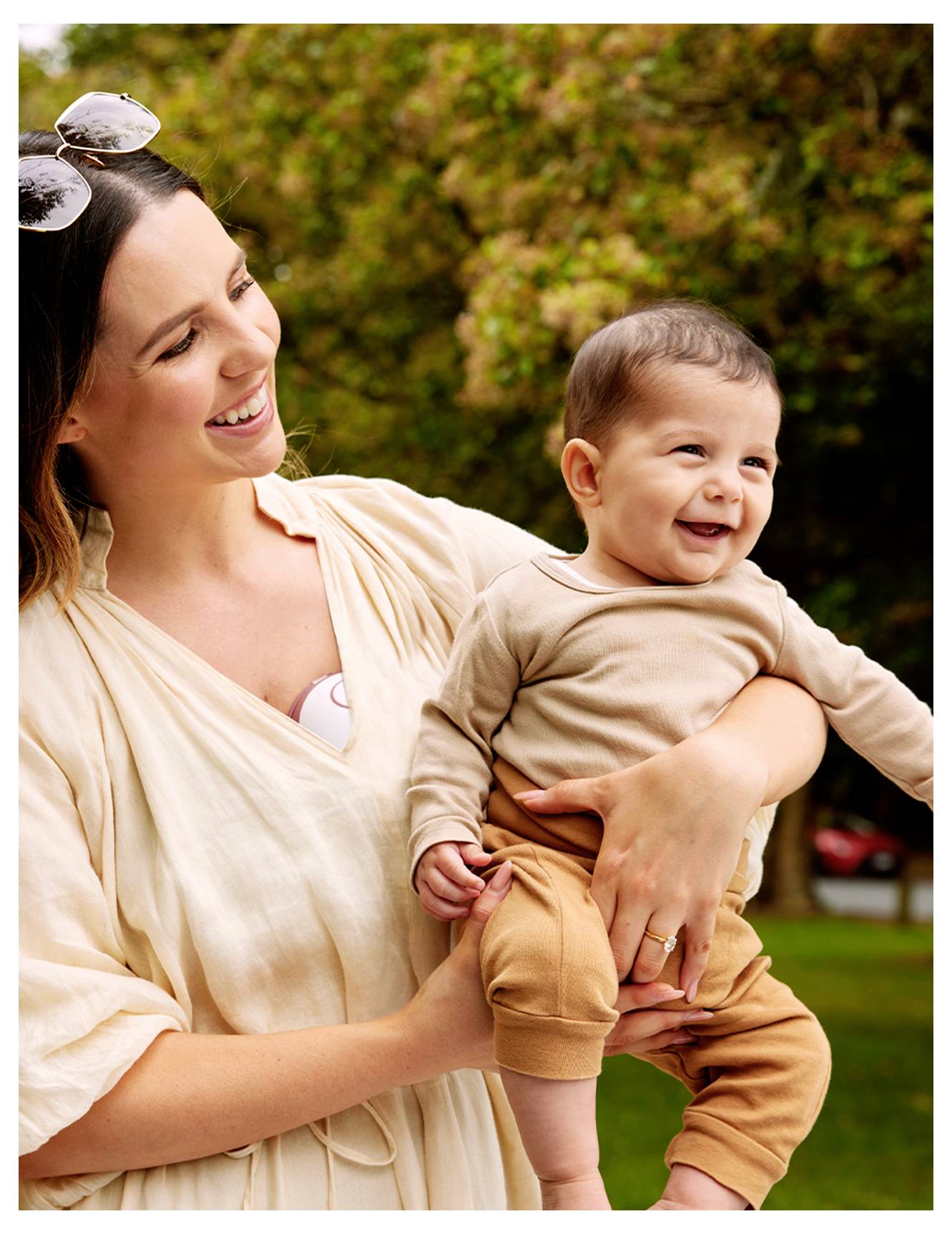 Una donna sorridente all'aperto che tiene in braccio un bambino allegro vestito in beige e marrone. Appaiono felici, con gli alberi sfocati sullo sfondo.