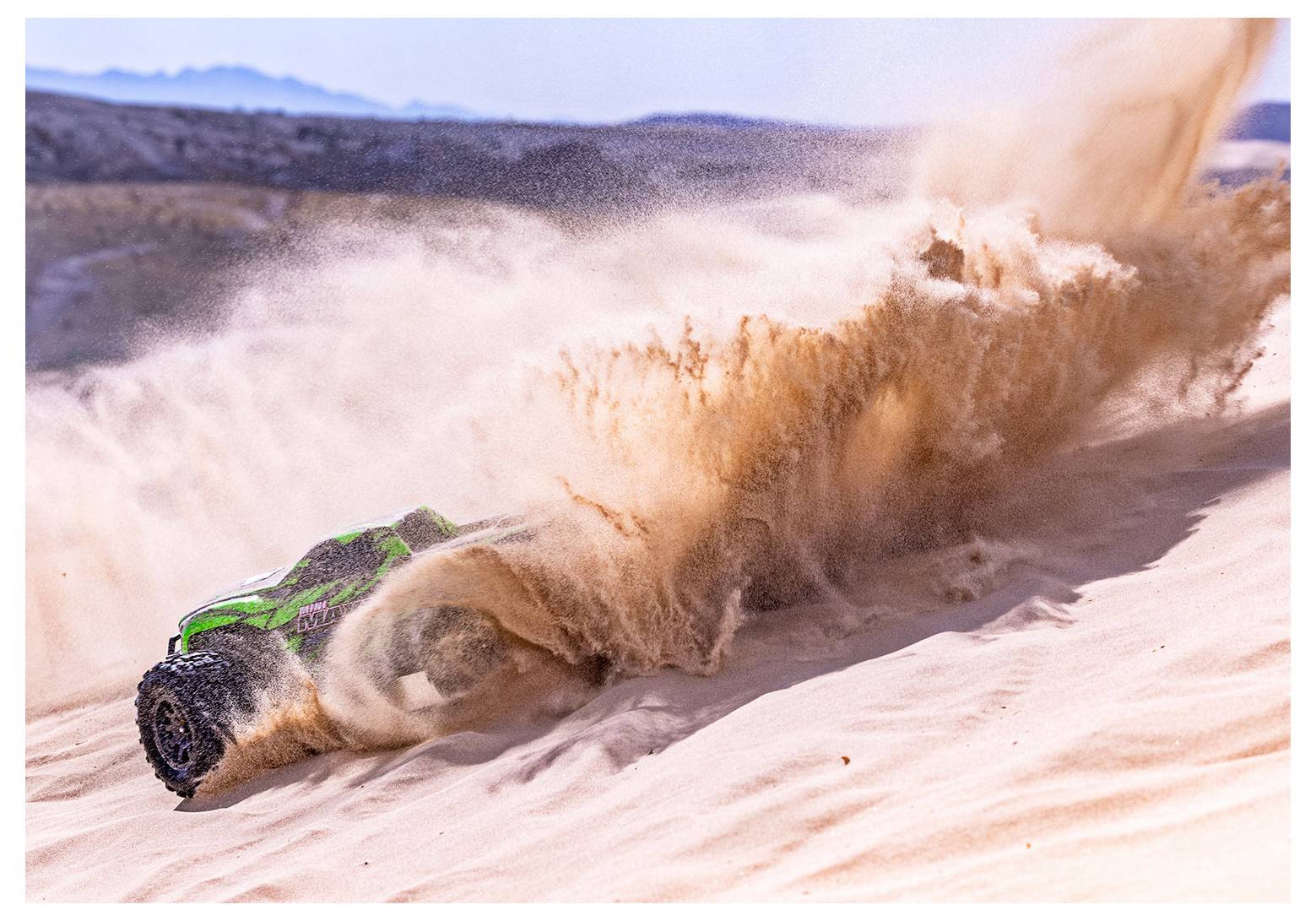 Un'auto radiocomandata verde solleva un'ampia scia di sabbia mentre sfreccia giù per una duna desertica, con colline visibili sullo sfondo.