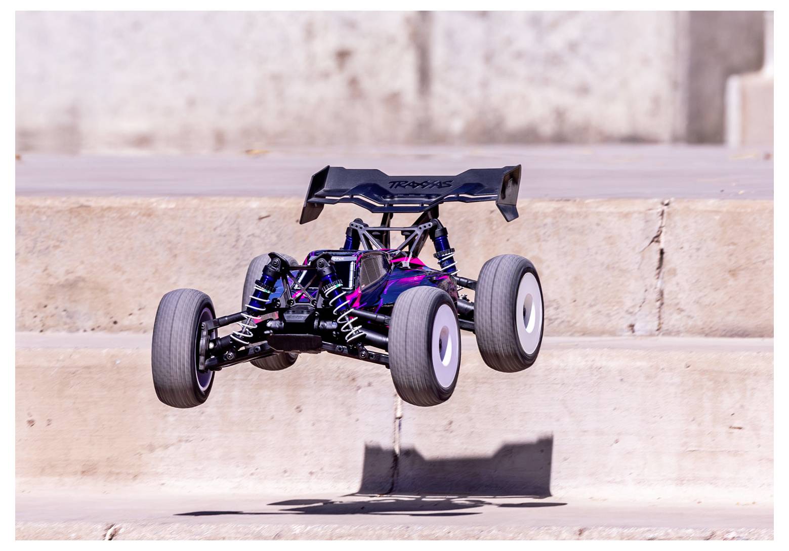 Un'auto radiocomandata con ruote grandi è in aria, saltando sopra gradini di cemento all'aperto. Il veicolo ha un design viola e nero.