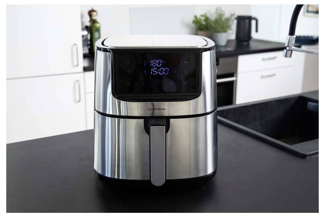 Un'air fryer in acciaio inossidabile su un bancone della cucina con display digitale che mostra '180°C'. Utensili da cucina e piante sullo sfondo.