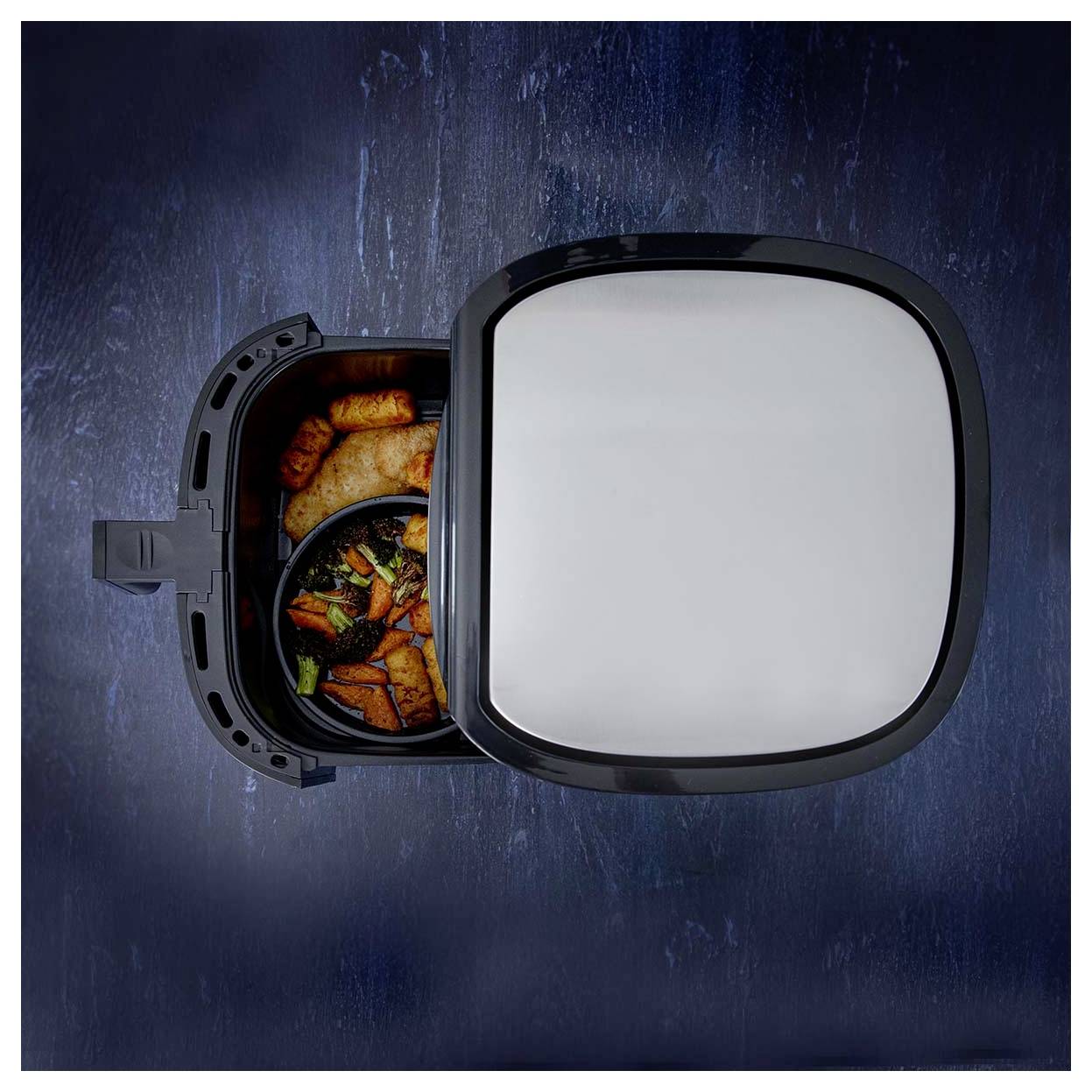 Cestello di friggitrice ad aria con pollo e verdure cotte, ripreso dall'alto, con un coperchio chiuso vicino su uno sfondo blu scuro.